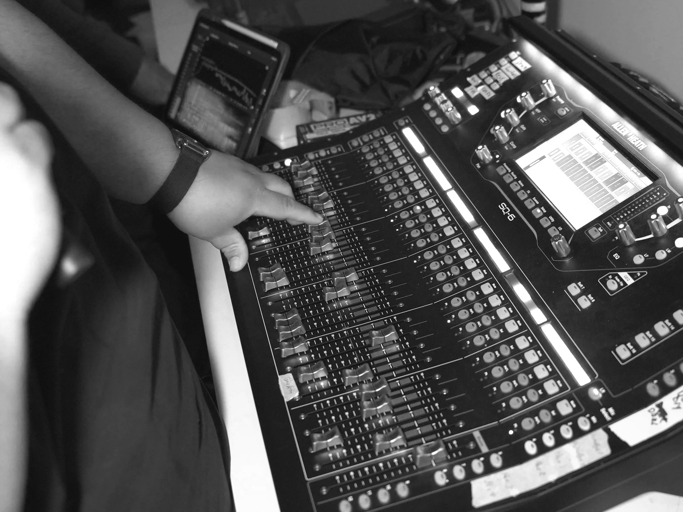 Black and white photo of a DJ's mixing console with an arm adjusting controls, accompanied by a smartphone showing audio waveforms.