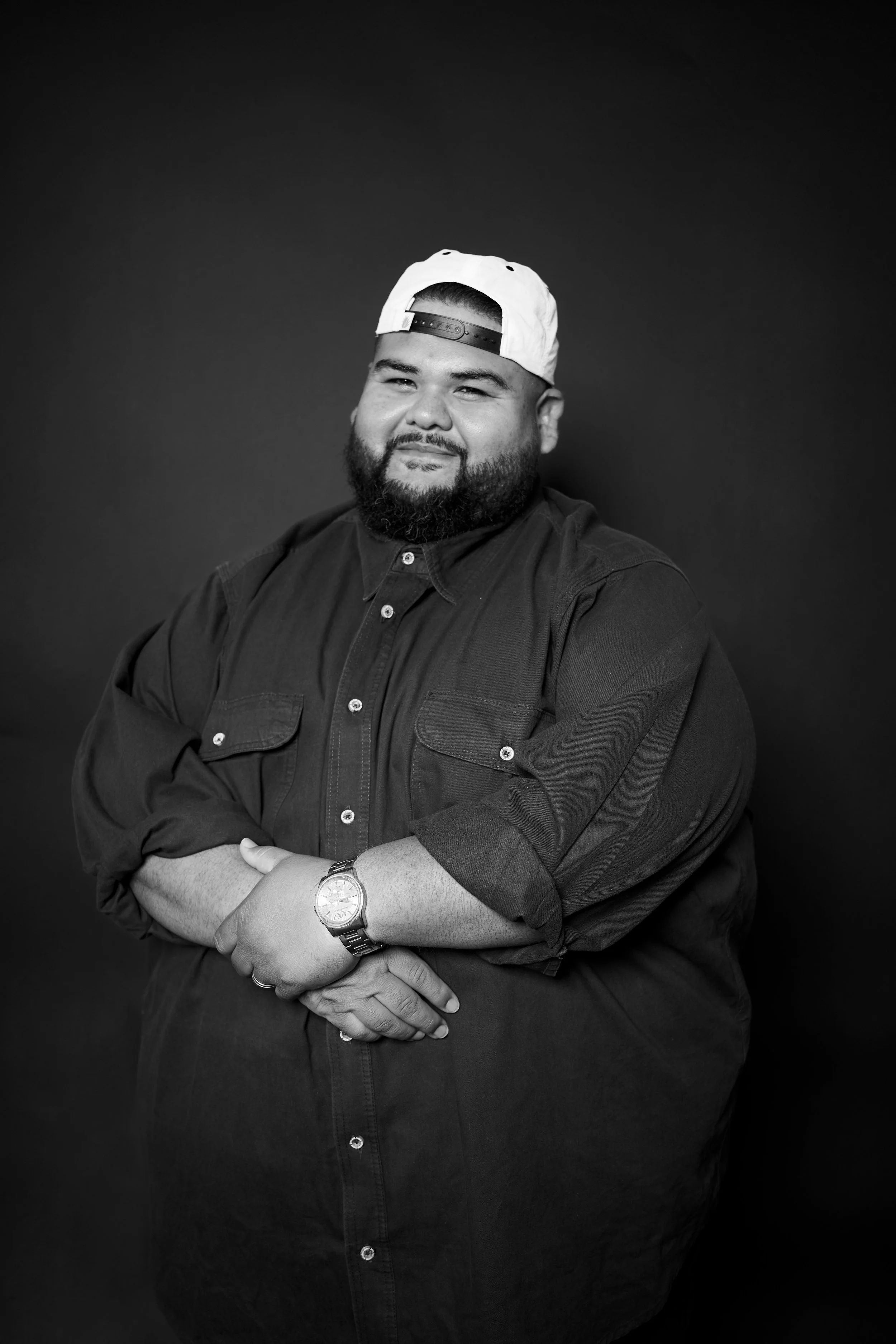 Black and white portrait of a man with a beard, wearing a backwards white baseball cap and a dark shirt, smiling and posing with crossed arms, against a dark background.