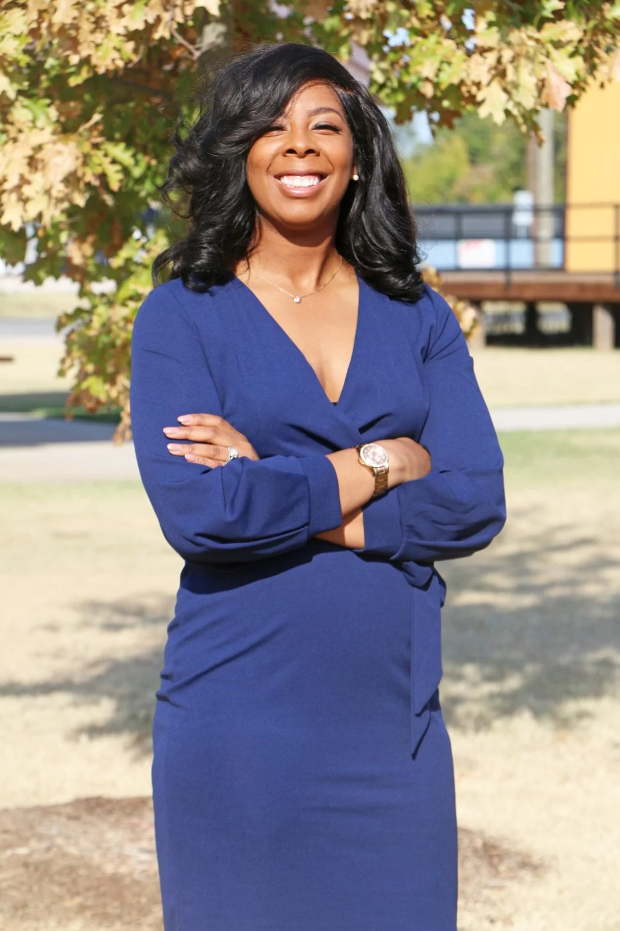 A smiling African American woman wearing a blue dress, standing outdoors with crossed arms, in front of a tree and building.