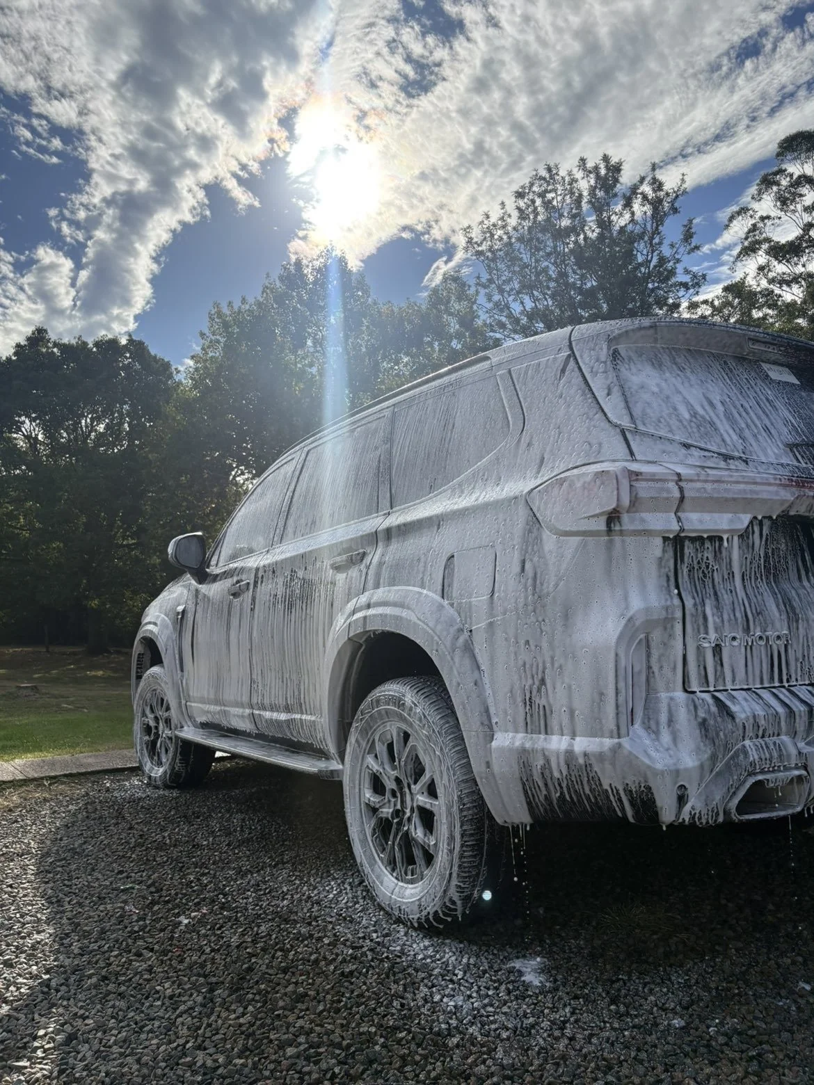 A silver SUV covered in soap suds during a car wash, with sunlight and clouds in the sky and trees in the background.