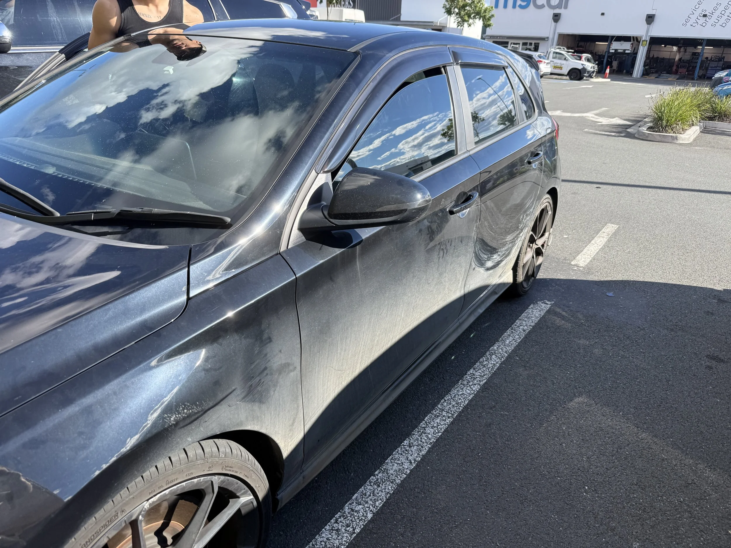 A close-up view of a black hatchback car parked in a parking lot, with clouds reflecting on its windows and body. Part of a person standing next to the car is visible in the background. The parking lot has white painted lines and nearby greenery.