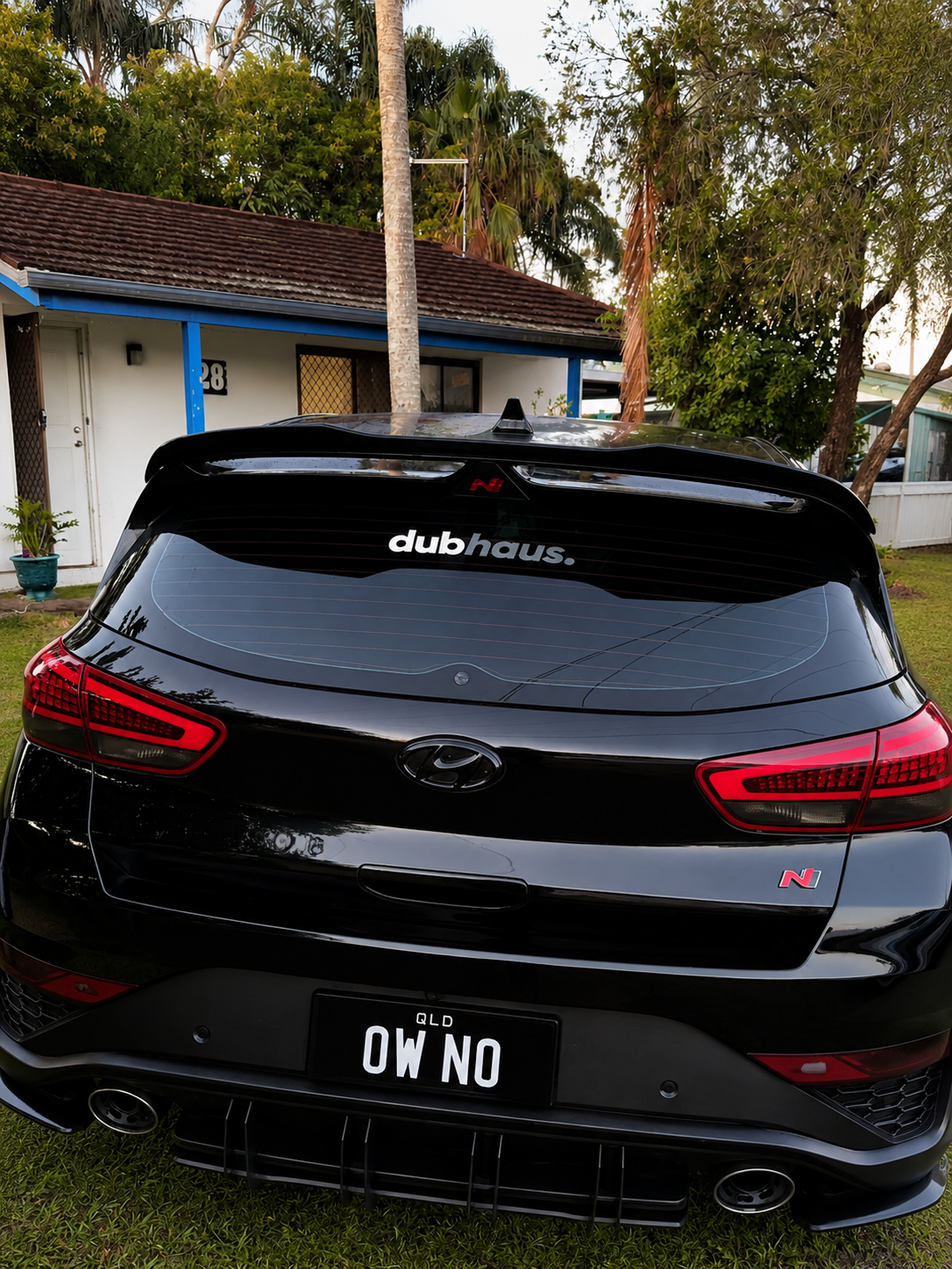 Black sedan with Australian license plate reading 'OW NO', parked in front of a house with a white wall and blue trim, surrounded by trees and plants.