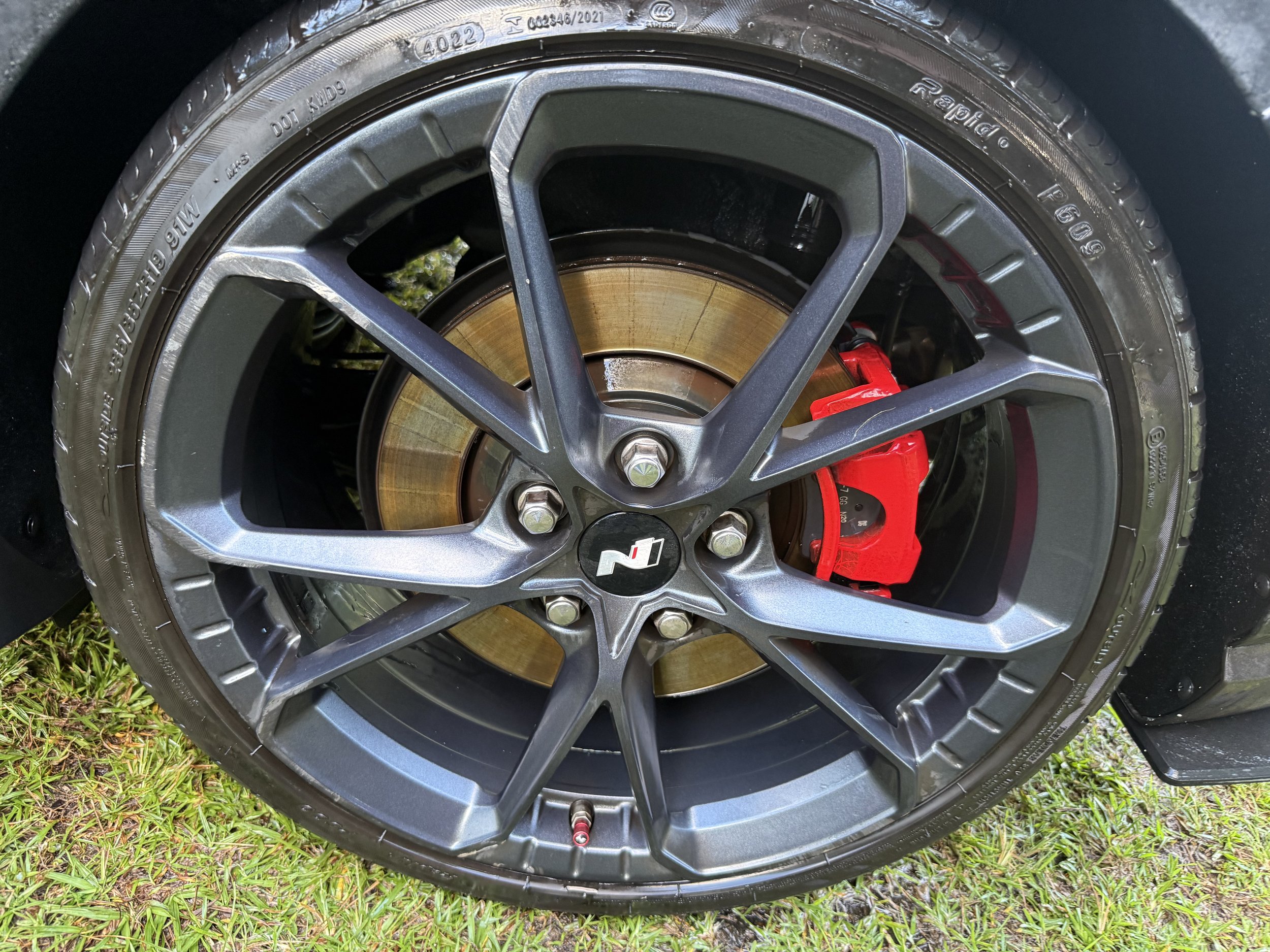 A car wheel with a black rim, red brake caliper, and a silver brake disc, sitting on grass and pavement.