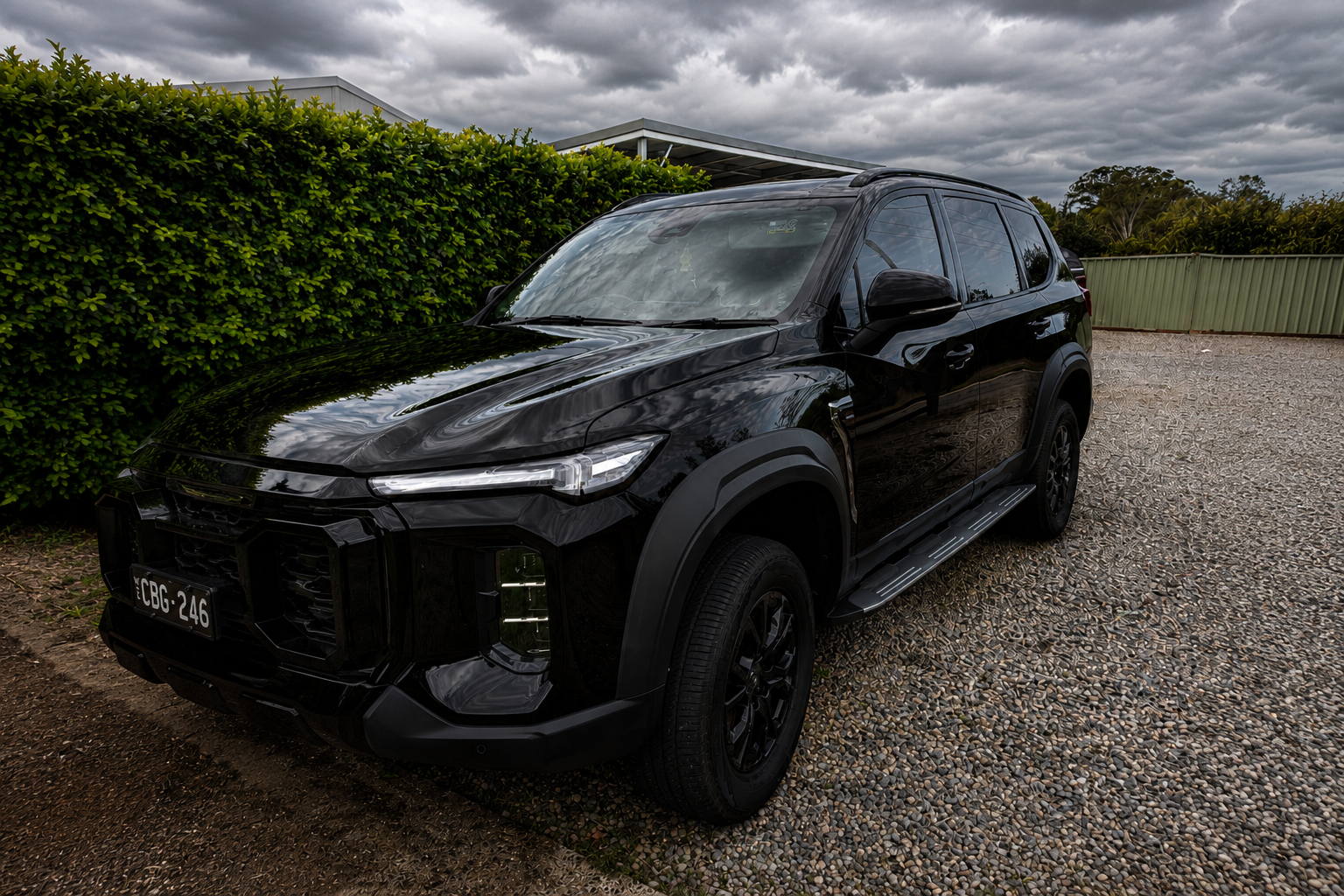 Black SUV parked on gravel driveway beside green hedge, cloudy sky overhead.
