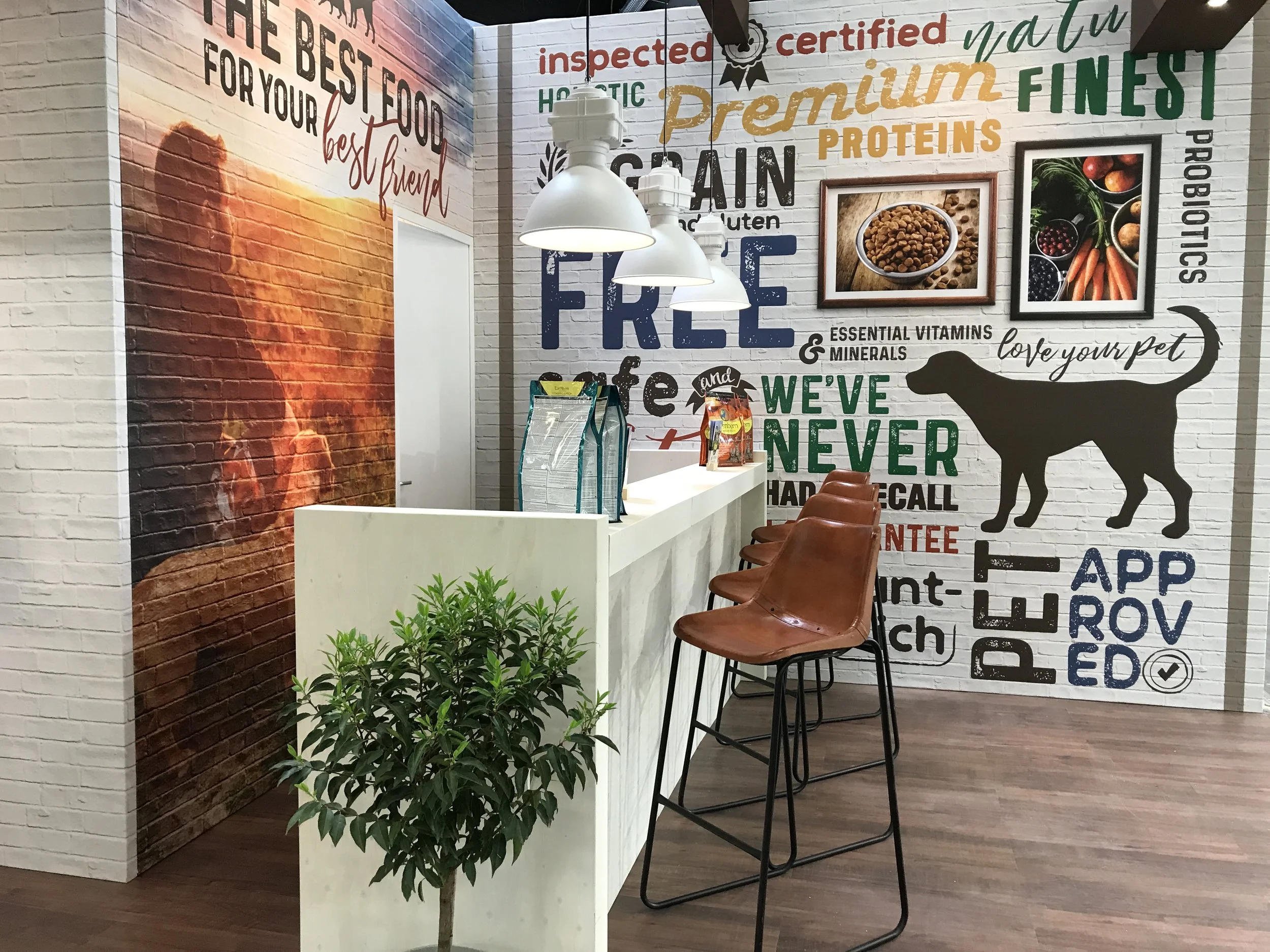 Interior of a health food or supplement store with white brick walls covered in colorful text and images. There's a row of four brown chairs along a bar-height counter, with a potted plant in front. The walls display words like 'organic,' 'frozen,' 'pet,' and pictures of different foods and supplements.