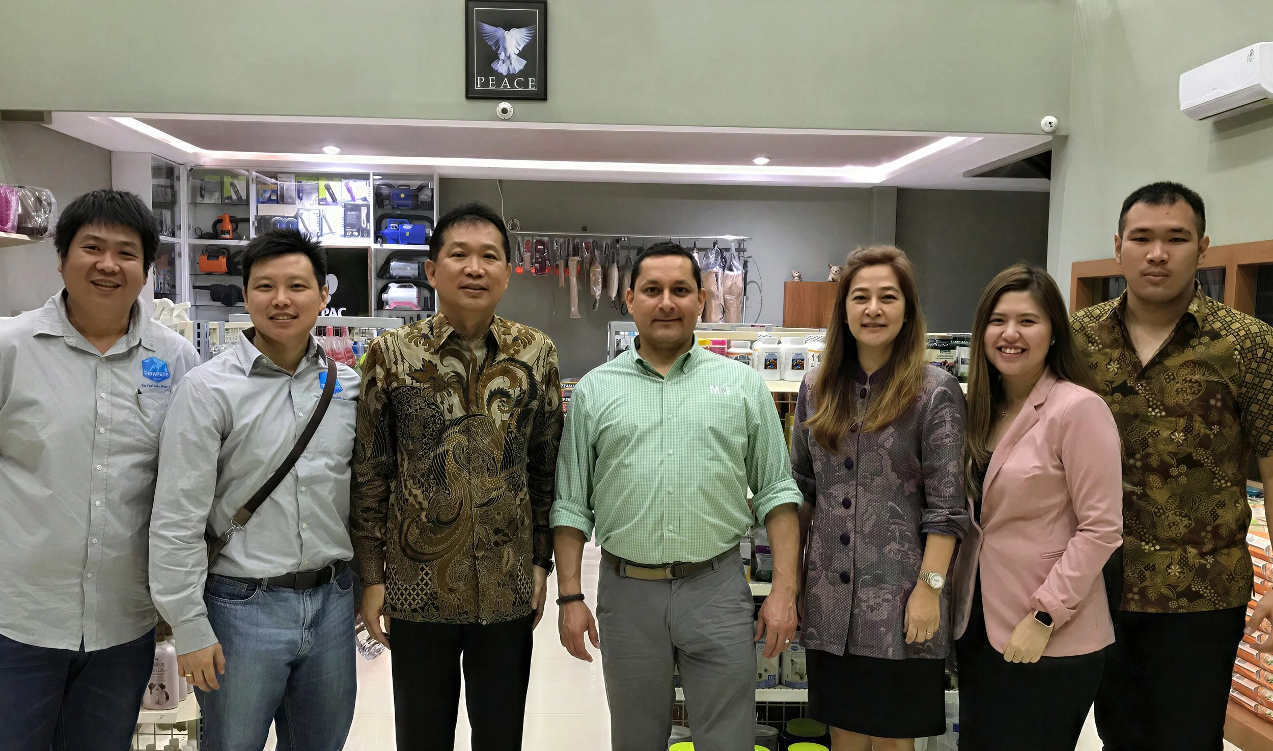Group of eight people standing side by side in a store with shelves of various products in the background, some wearing patterned shirts and others in plain attire.