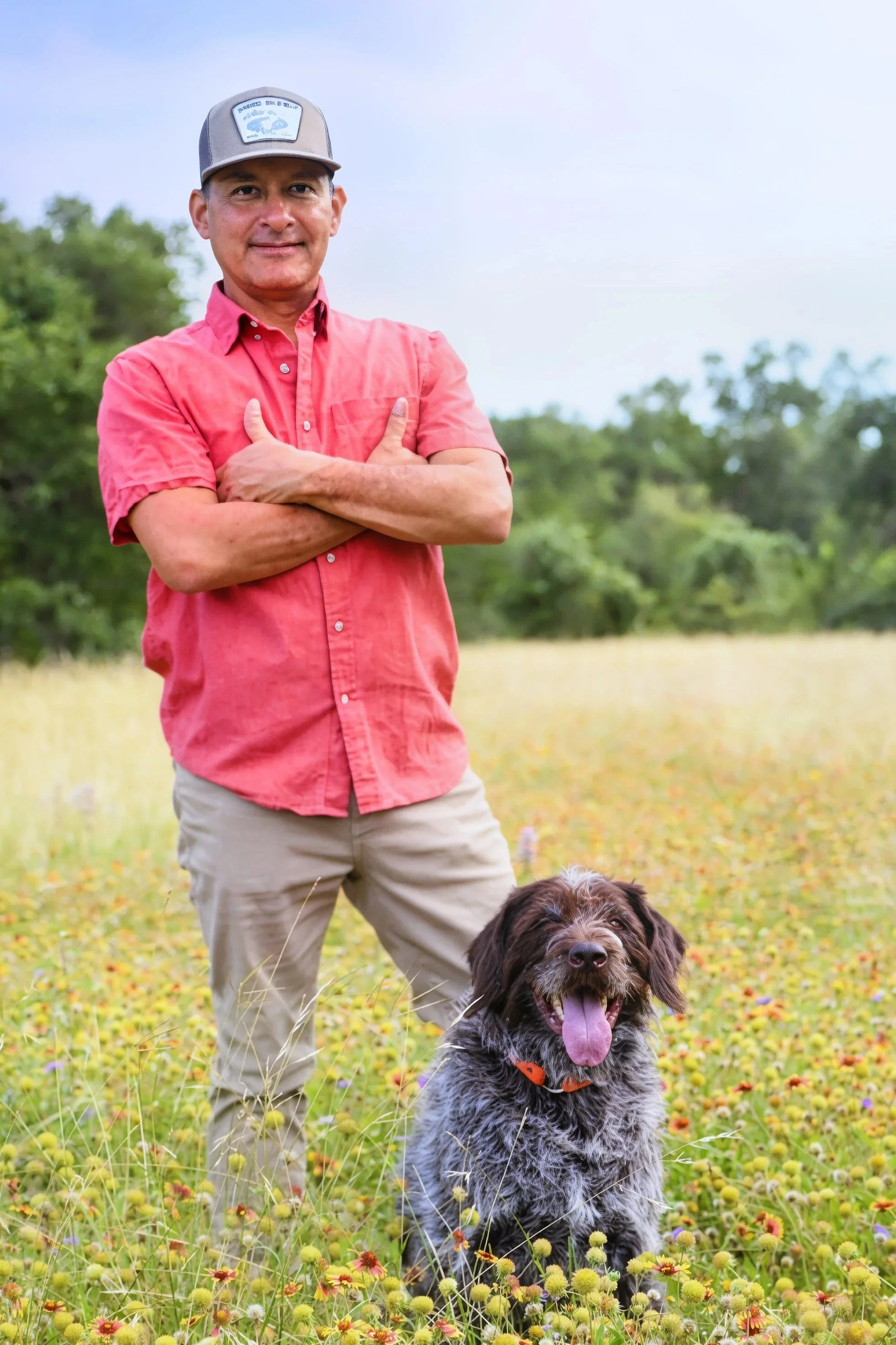 A man in a red shirt and beige pants standing in a field of yellow and purple flowers with a dog sitting in front of him. The man is smiling and has his arms crossed, wearing a gray cap. The dog is a large, fluffy breed with a thick coat, tongue hanging out, and an orange collar. Trees are visible in the background under a blue sky.