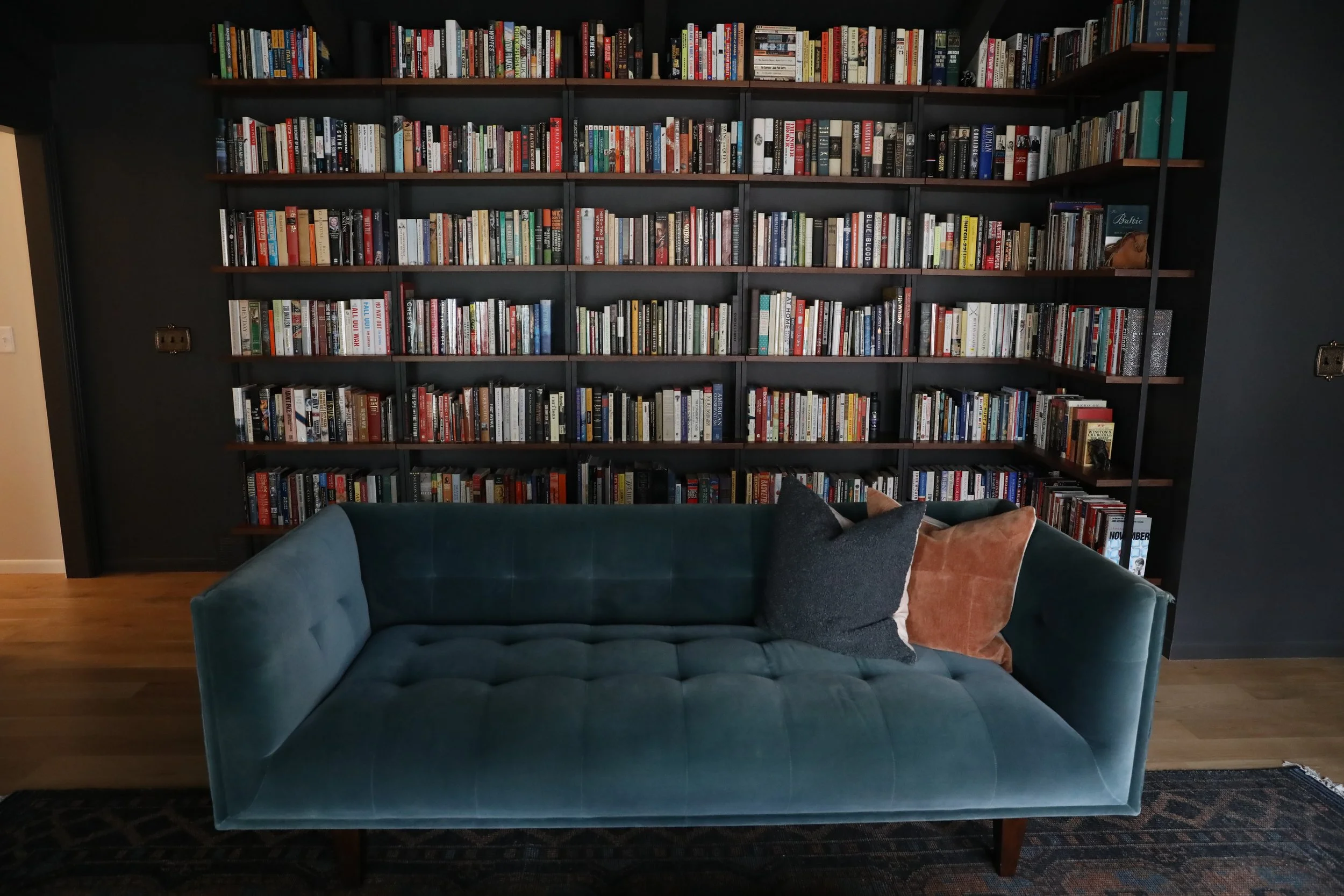 A view of a large bookshelf filled with books against a dark wall with a wooden floor. In front, there is a blue velvet tufted sofa with two pillows, one dark gray and one beige. Part of an area rug is visible beneath the sofa.