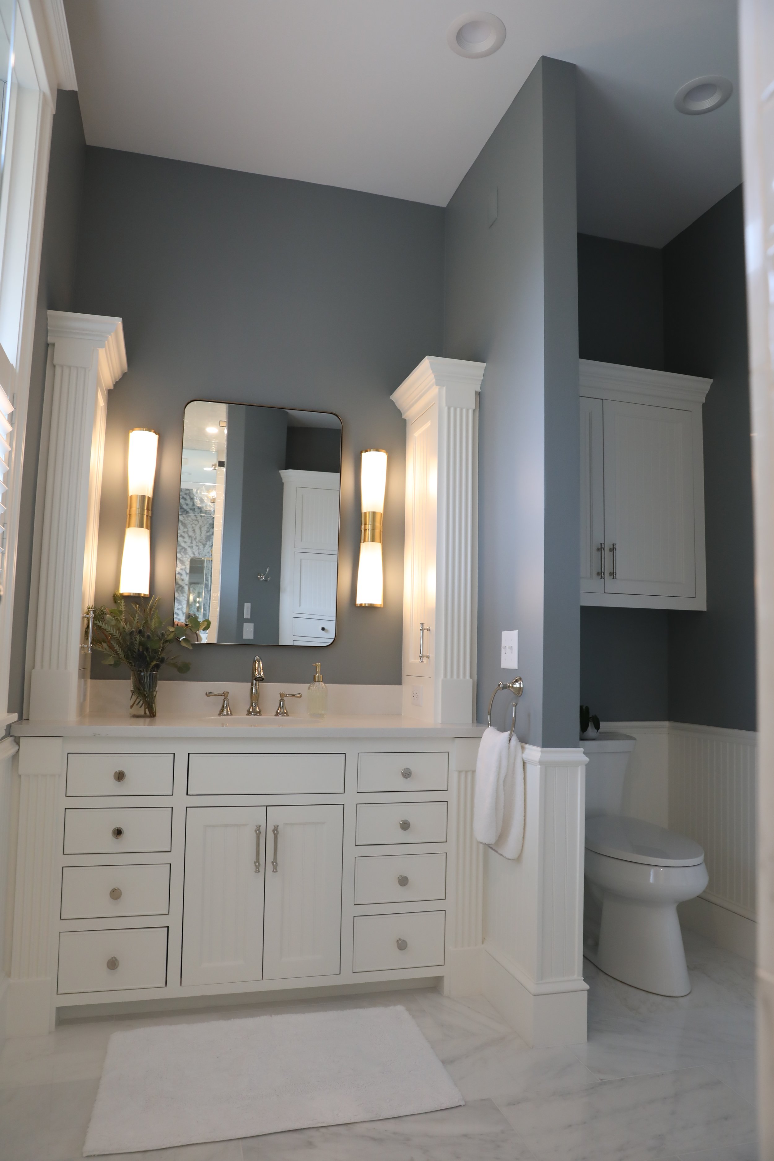 A modern bathroom with a white vanity, framed mirror, wall-mounted light fixtures, a vase with flowers, a white towel hanging on a silver hook, a toilet, white cabinetry, and gray walls.