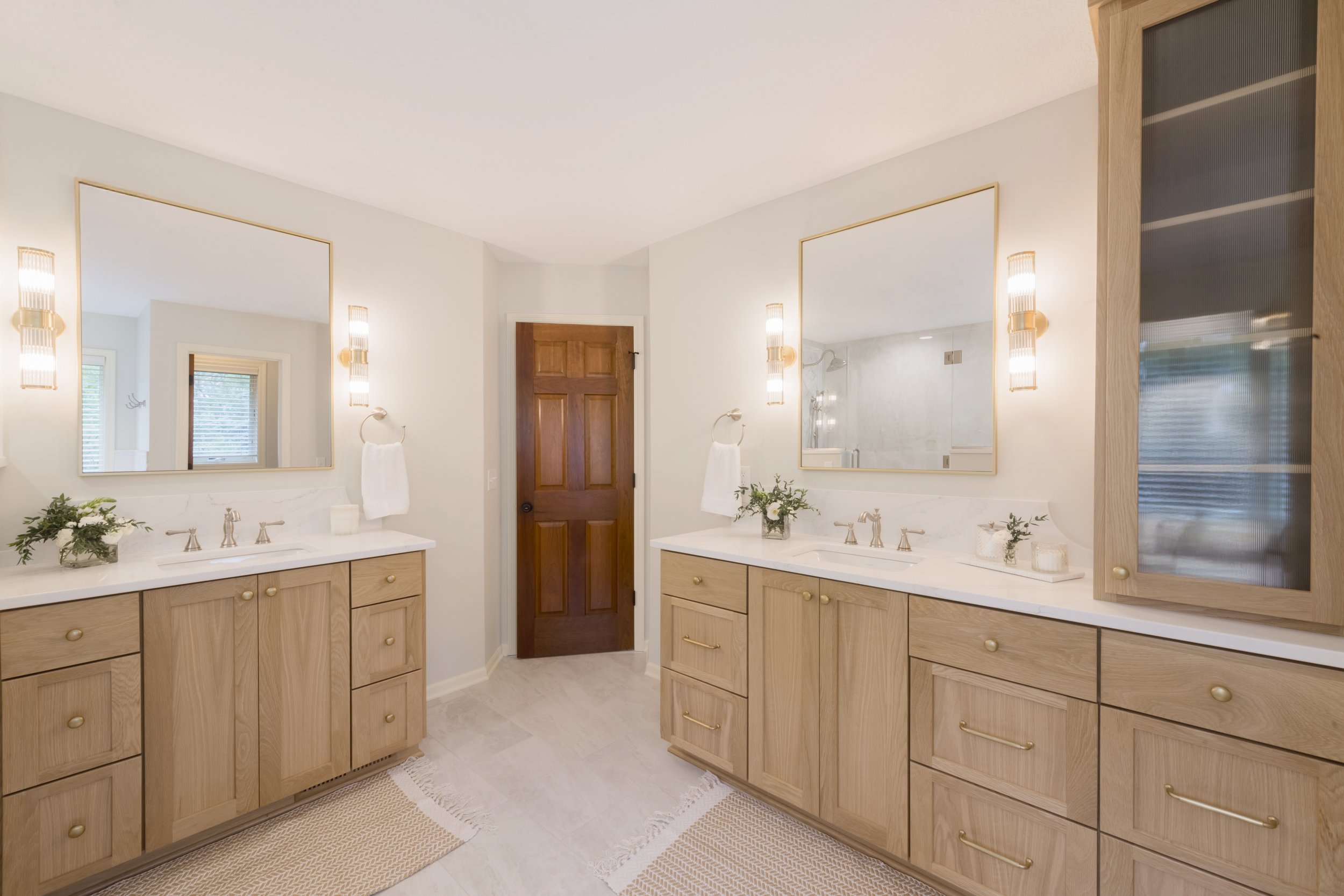 Bathroom with double vanity, large mirrors, wooden cabinets, and a door leading to a shower area. Decor includes potted plants, towel holders, and wall sconces.