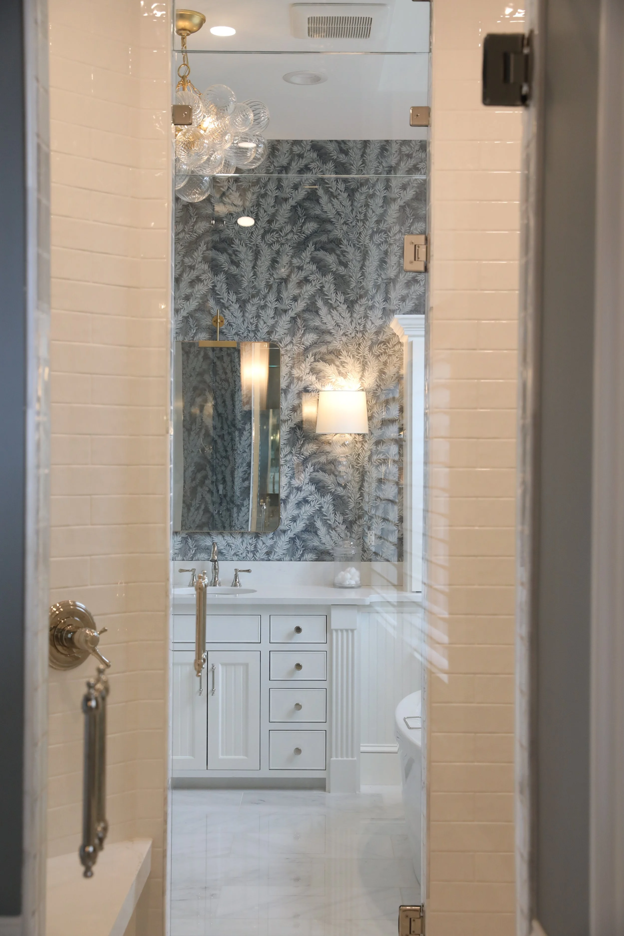 A view into a bathroom from an adjacent room. The bathroom has a white vanity with a mirror, a wall-mounted light fixture, a patterned wallpaper with a botanical design, a wall lamp, a shower area, and a visible toilet in the corner.
