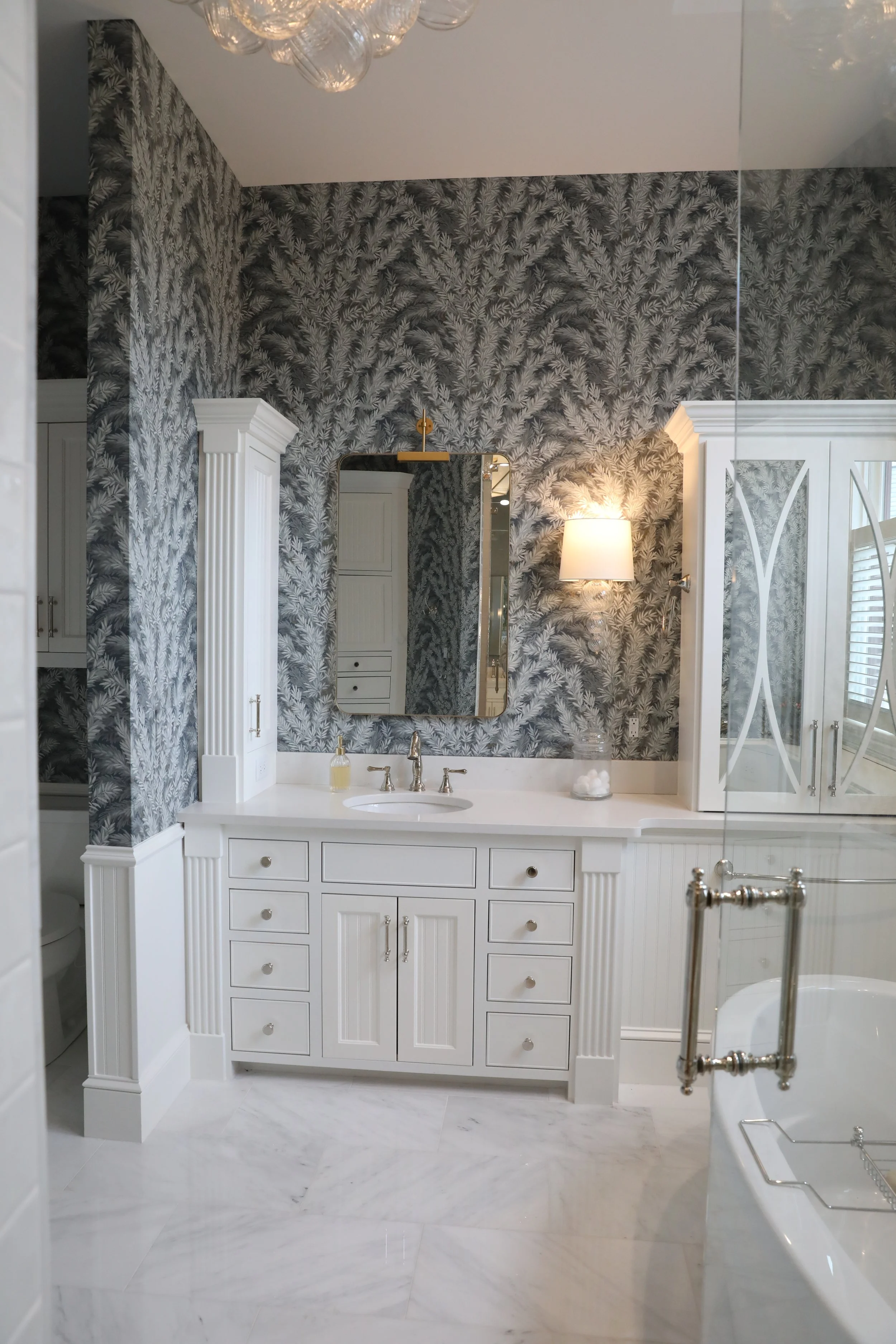 A bathroom with a white vanity, a mirror, and a wall-mounted lamp. The walls are decorated with a gray leafy pattern, and there is a large mirror and glass shower door reflecting part of the room. The floor is made of white marble tiles.