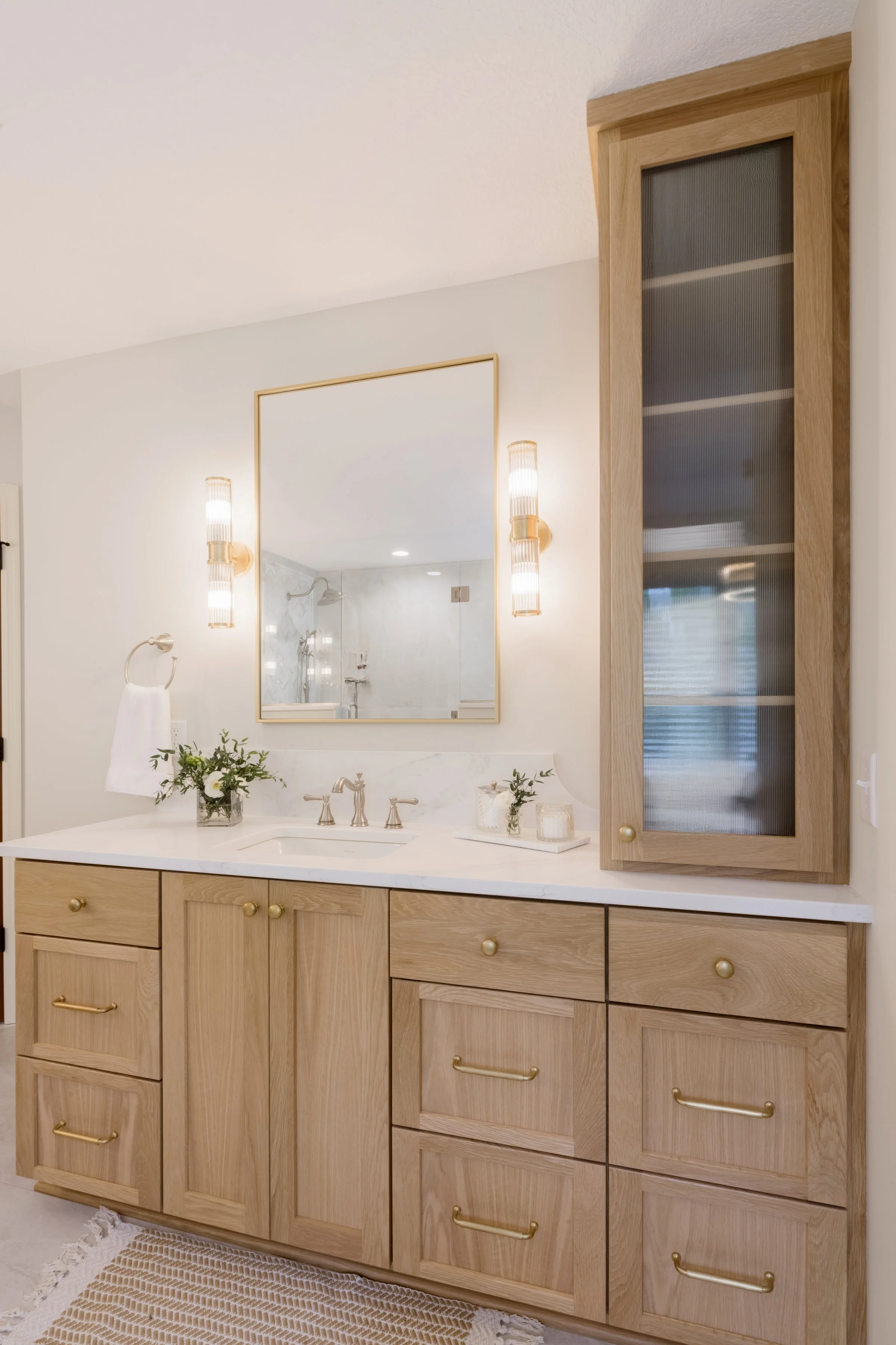 Bathroom vanity with wooden cabinets, a large mirror, two wall-mounted light fixtures, a vase with flowers, and a cabinet with a glass door.