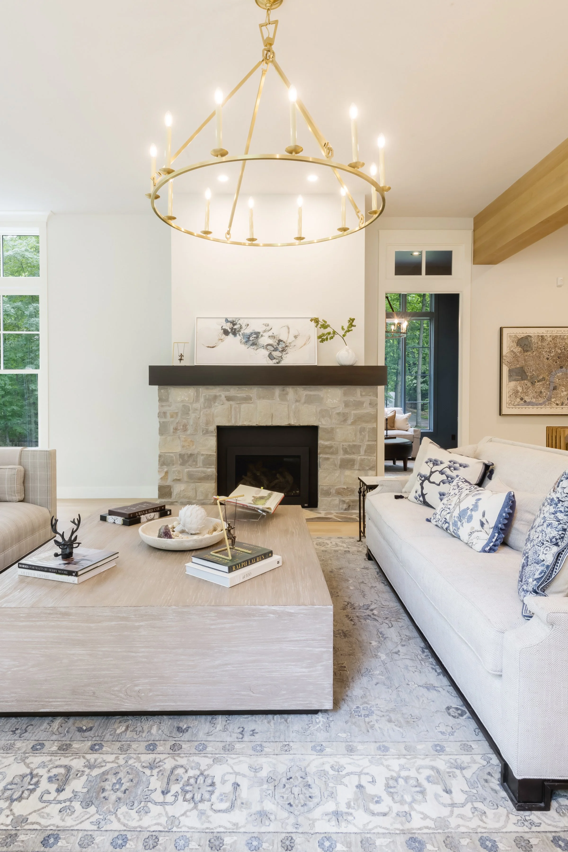 Living room with a stone fireplace, a large wooden coffee table with books and decorative items, a white sofa with patterned pillows, a chandelier, large windows, and artwork on the walls.
