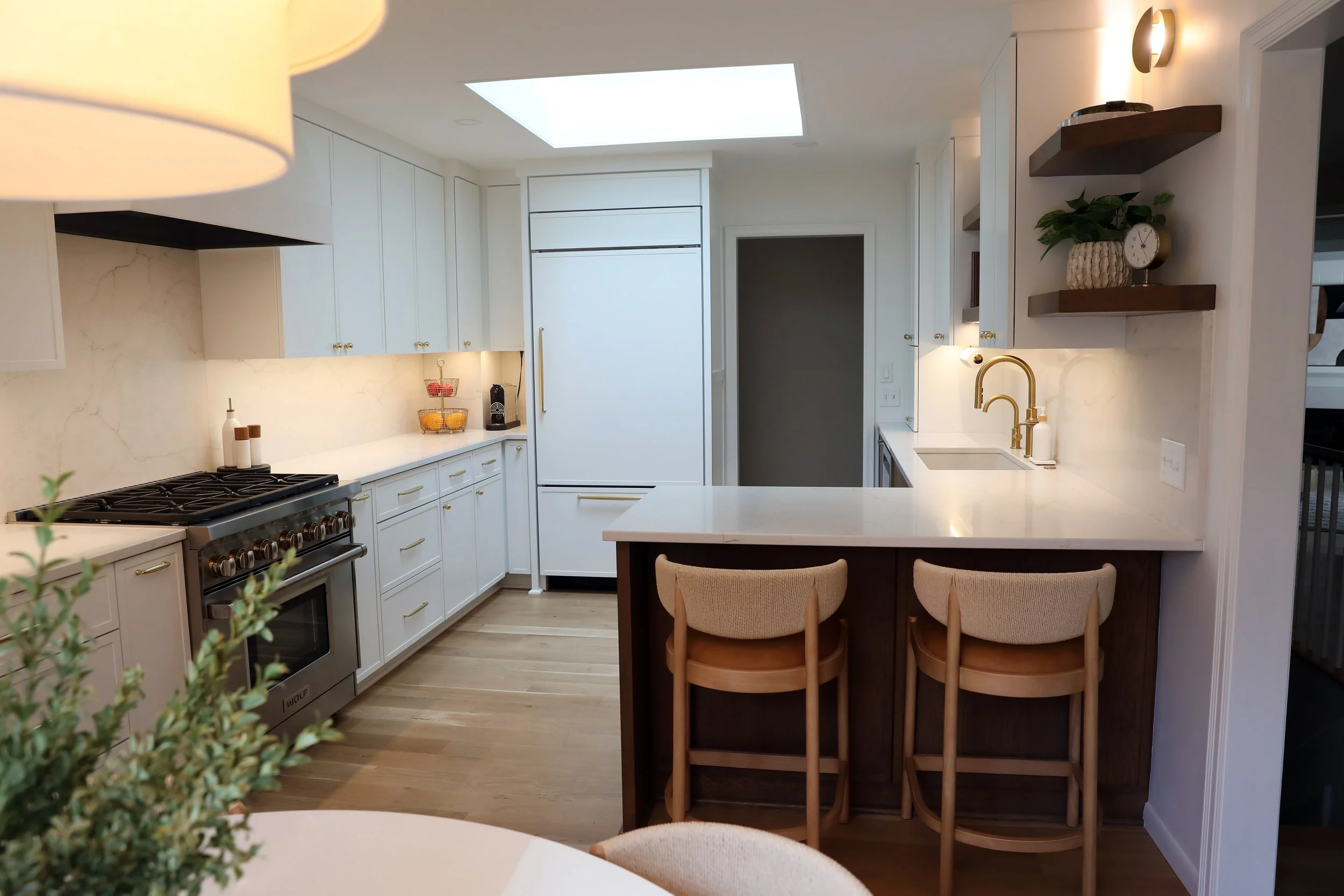 Modern kitchen with white cabinets, marble backsplash, gold hardware, and a breakfast bar with three chairs. Skylight and wall-mounted shelves with decor, plant, and clock.