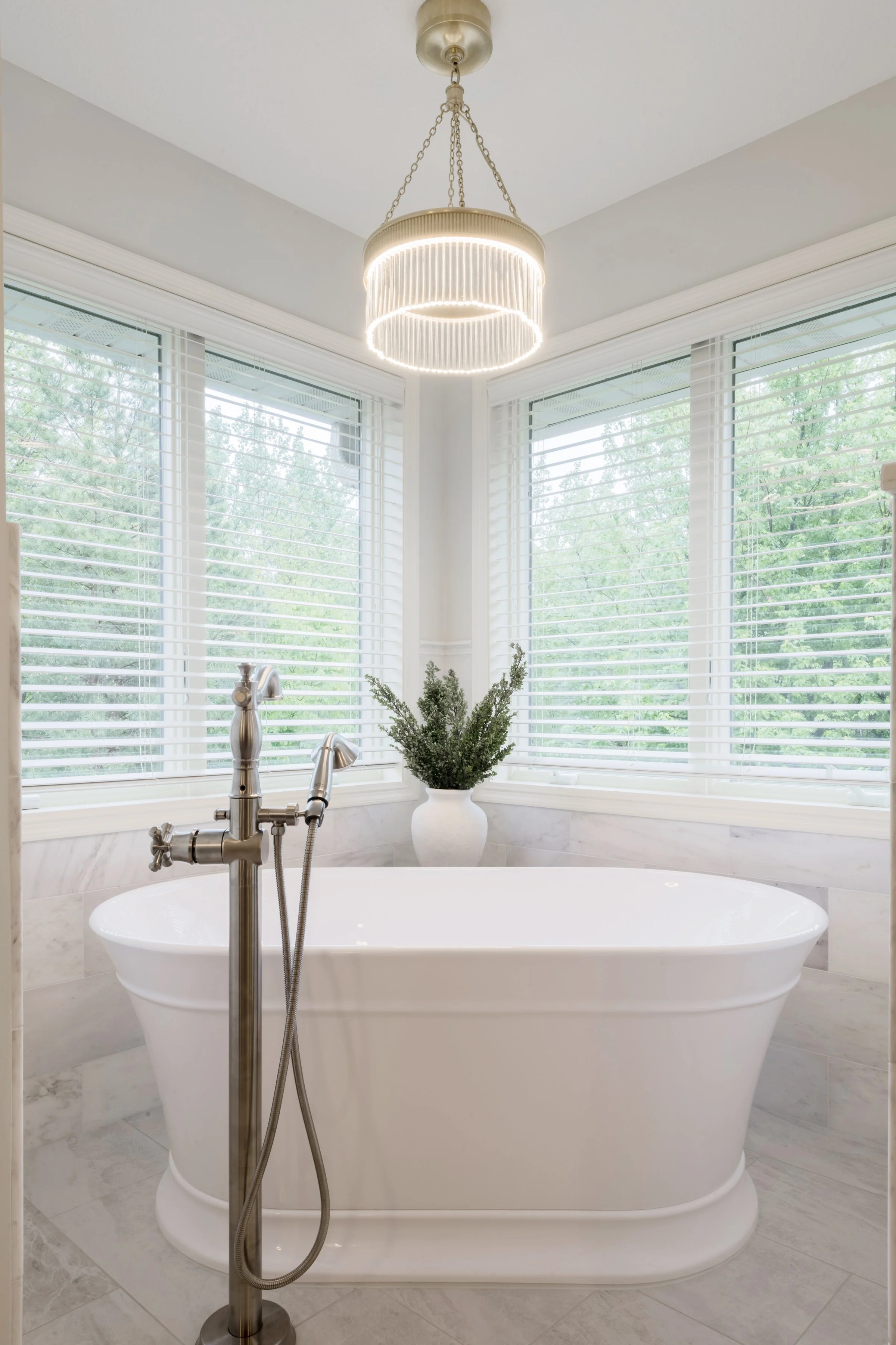 A white freestanding bathtub in a bright bathroom with large windows. There is a silver floor-mounted bathtub faucet and a green plant in a white vase on the windowsill. A modern chandelier hangs from the ceiling.