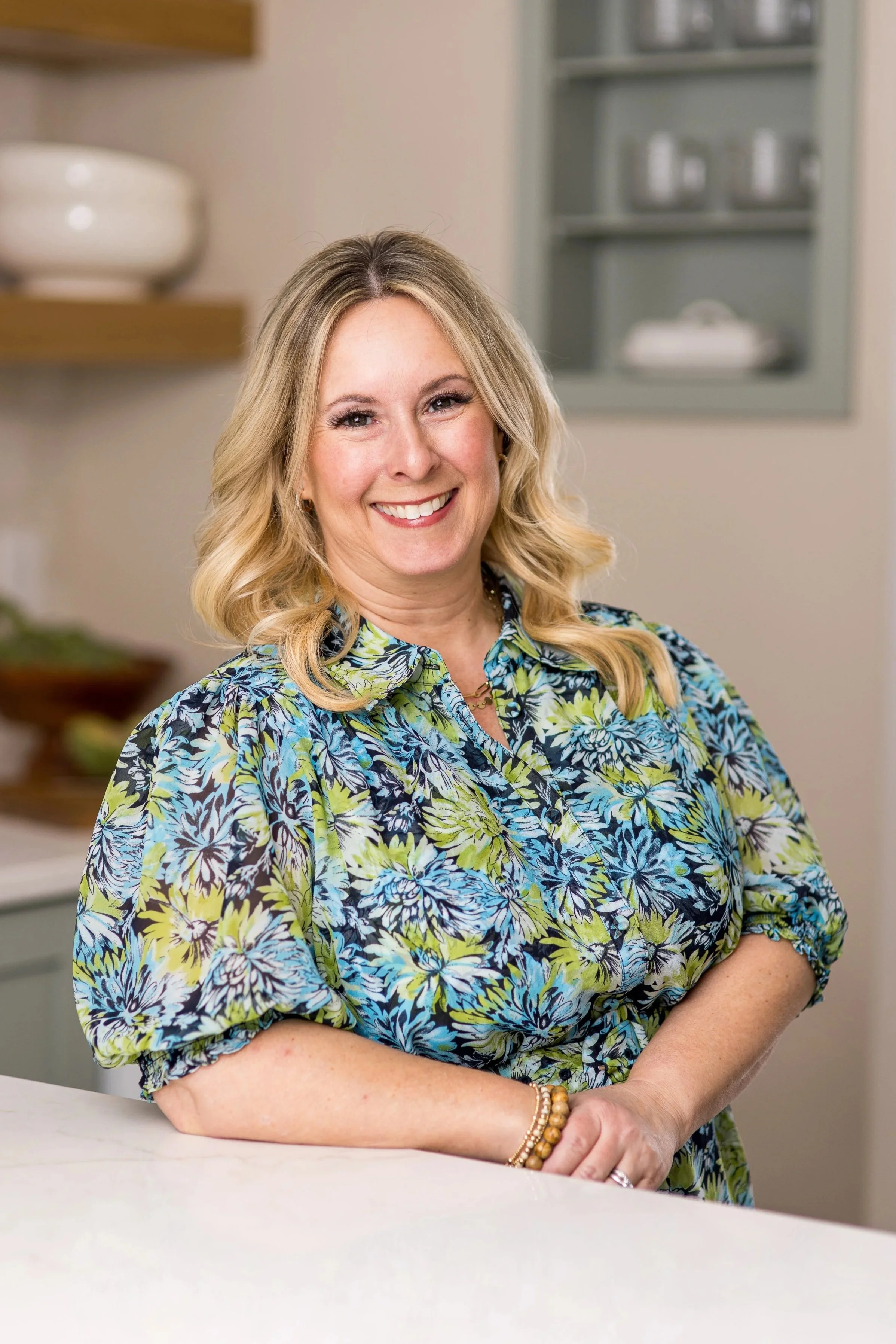 A woman with blonde wavy hair, wearing a colorful floral blouse, sitting at a white table in a kitchen, smiling at the camera.