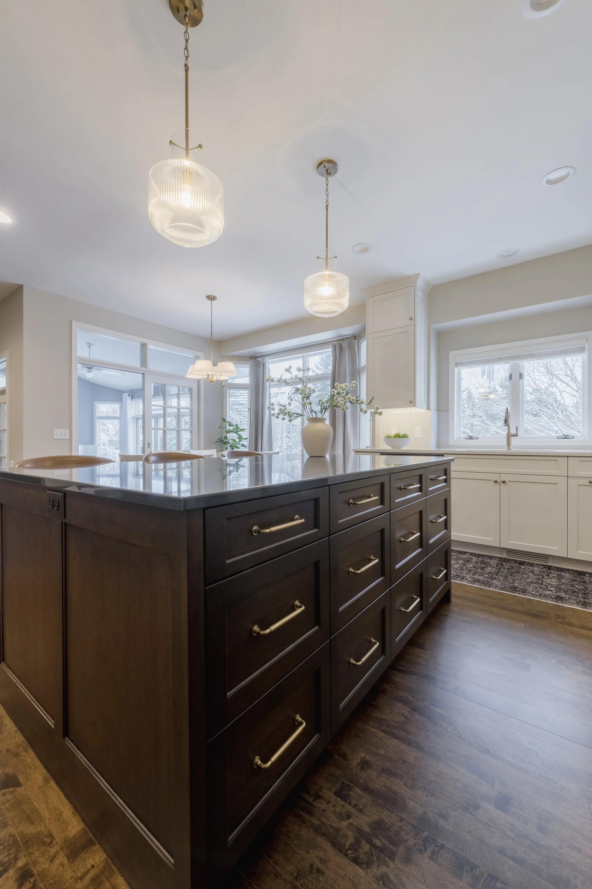 Kitchen with an island counter, dark wood cabinets, white upper cabinets, large windows, a flower vase on the island, and pendant ceiling lights.