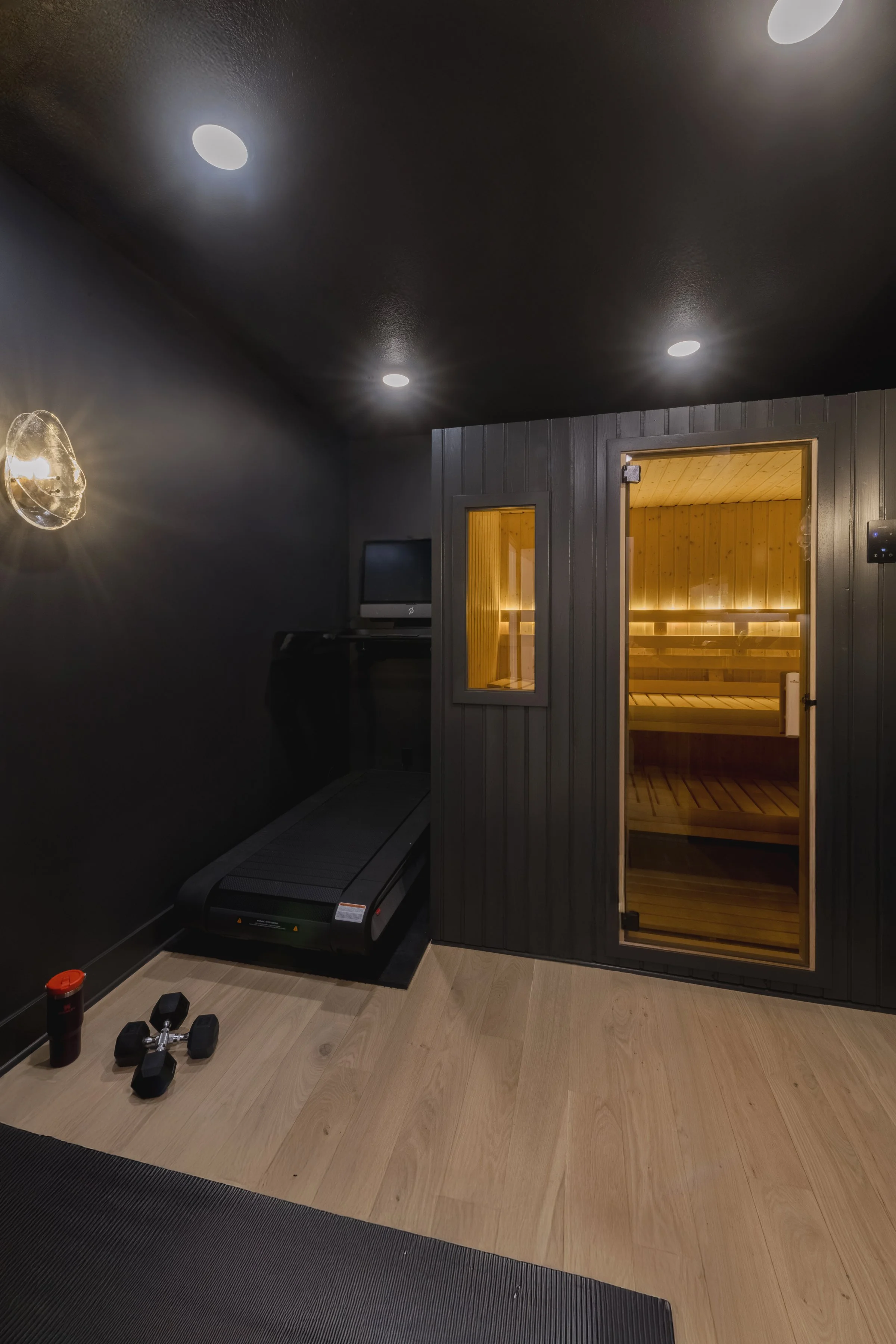 A modern room with black walls and ceiling, featuring ceiling lights, a treadmill, a small TV, and a sauna with wooden interior and glass door.