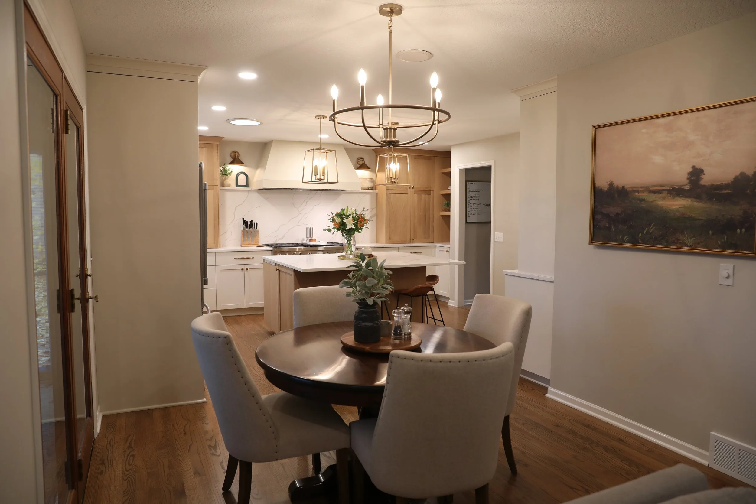 A view of a modern, cozy kitchen and dining area with a round dining table surrounded by beige upholstered chairs, a bouquet of flowers on the table, and a spacious kitchen with wooden cabinets, white marble backsplash, and pendant lighting fixtures.