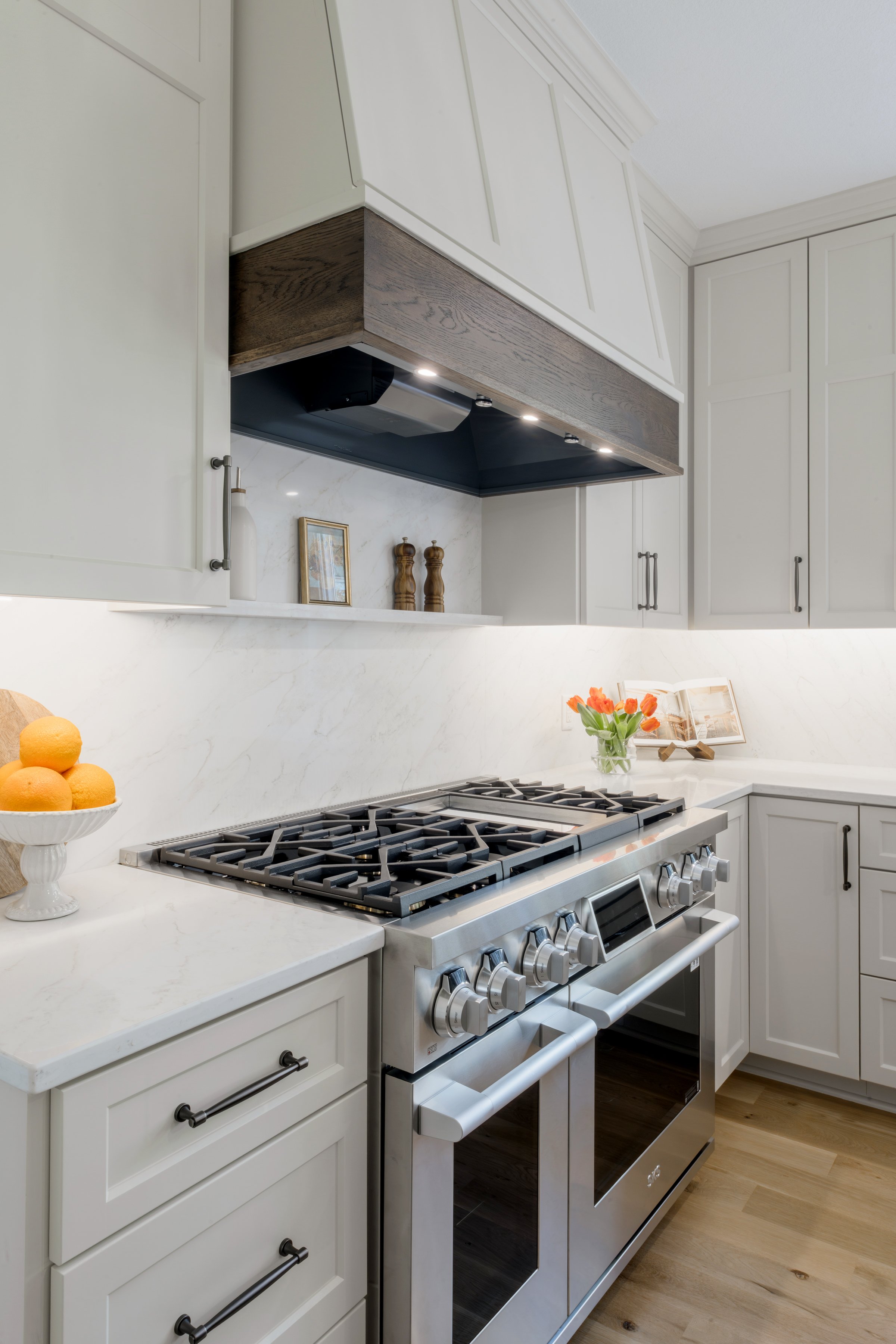 A modern kitchen with white cabinets, a stainless steel gas stove, a white marble countertop, a vase with orange tulips, and decorative items on the counter. There are wooden accents under the cabinets and a small framed picture.