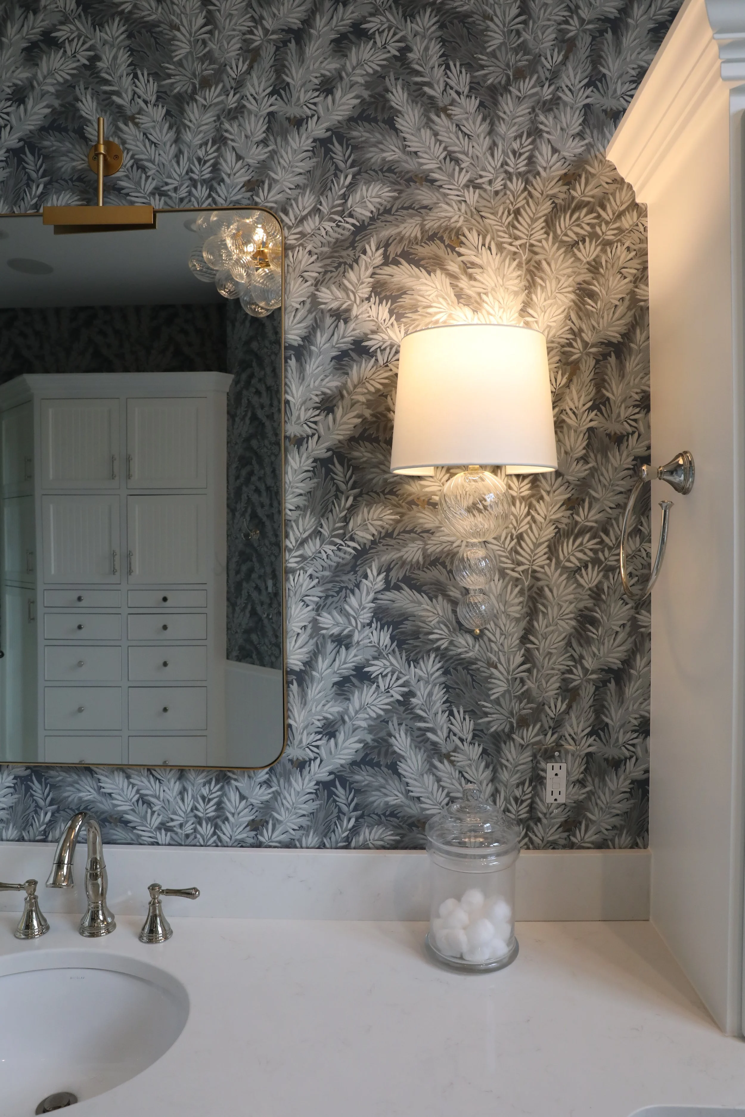 Bathroom vanity with a white countertop, a silver faucet with two handles, a large mirror, a wall-mounted lamp with a white shade, a decorative glass lamp with a clear base, a glass container filled with cotton balls, and a patterned wallpaper with g