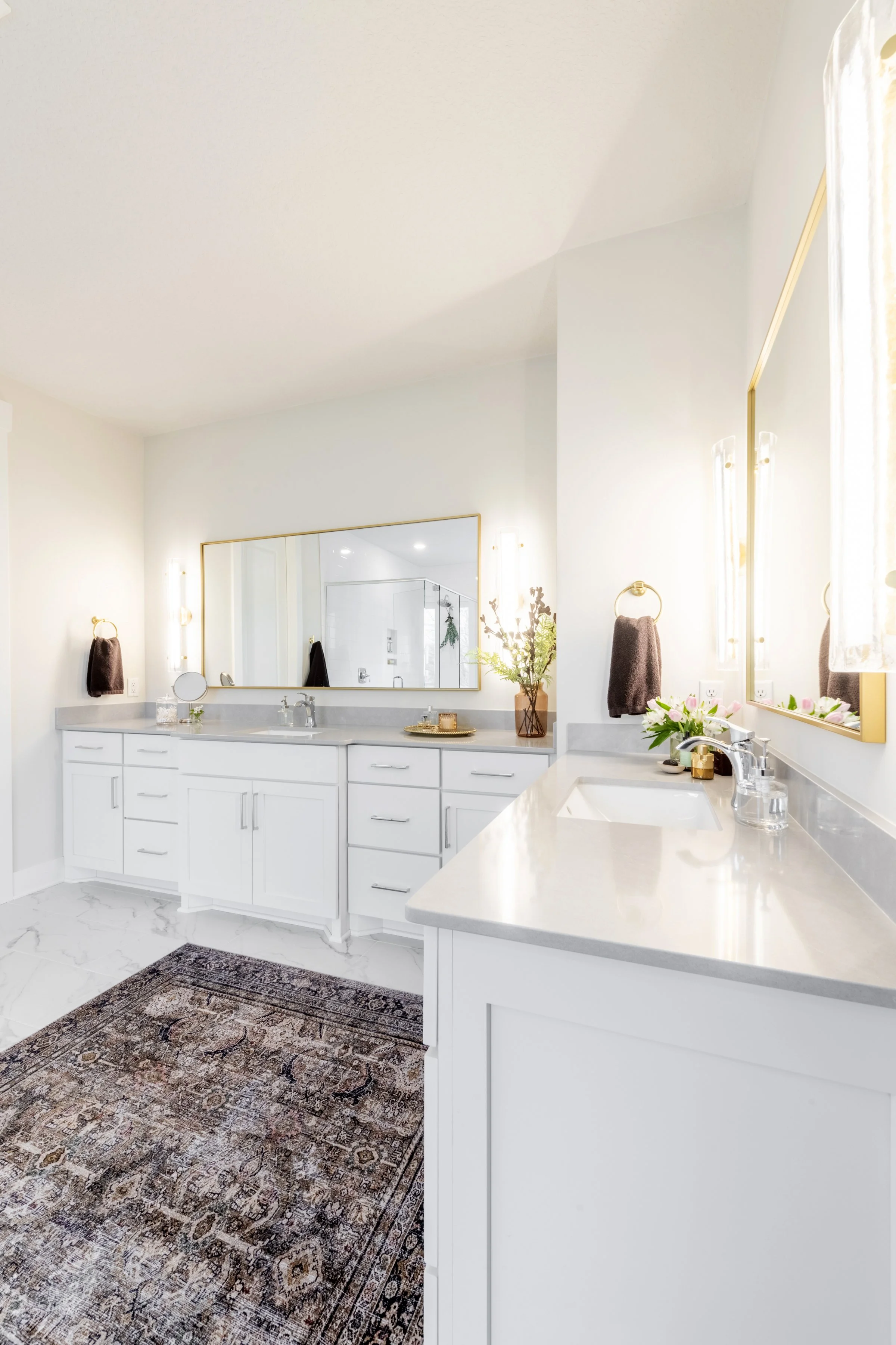 Bright bathroom with white cabinetry, large mirror, vase of flowers, towel rings with brown towels, and a colorful rug.