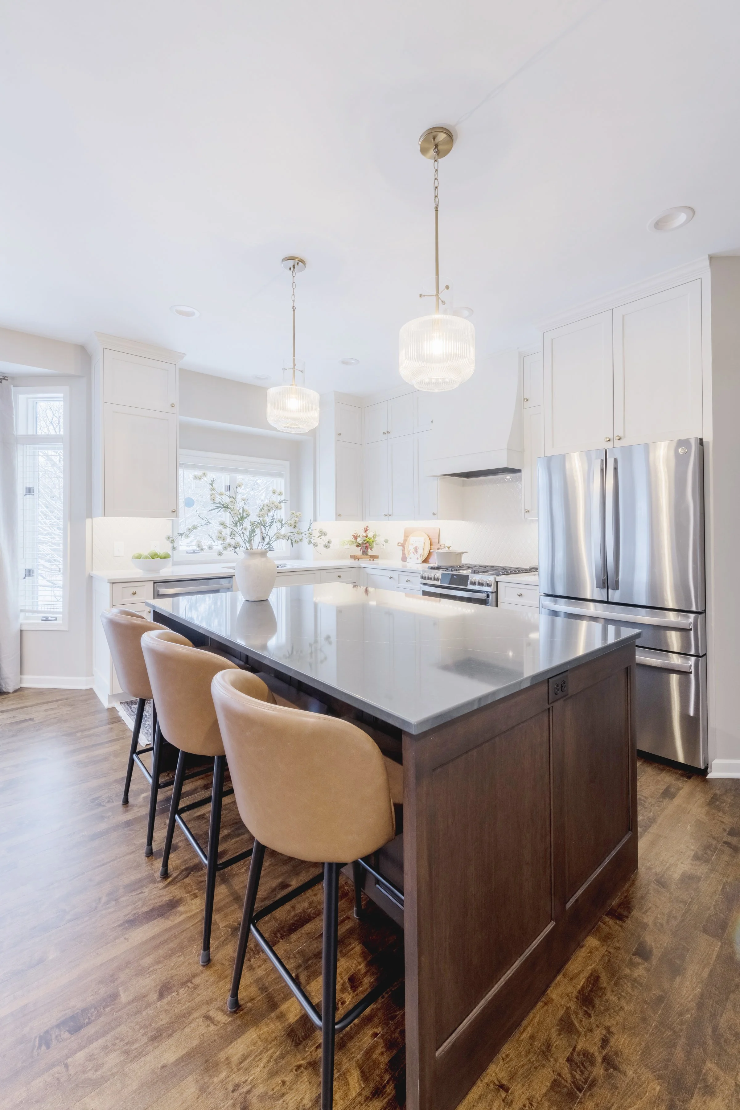 A modern white kitchen with a dark wood island, beige bar stools, white cabinetry, stainless steel refrigerator, and pendant lights.