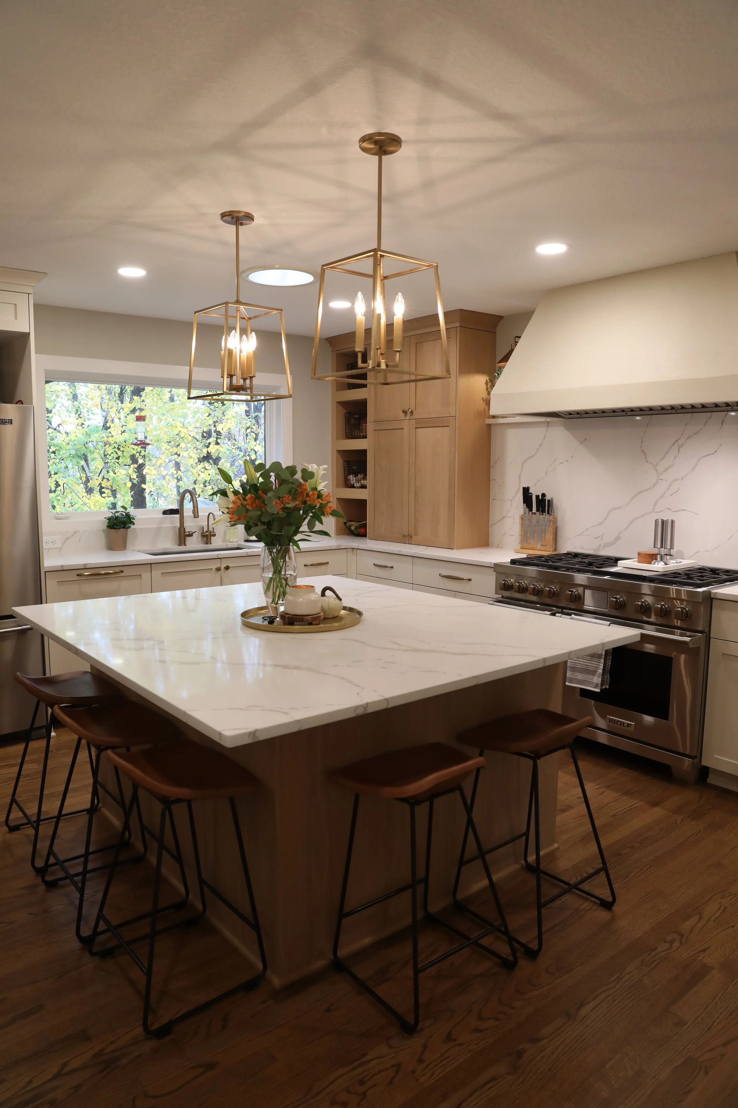 Modern kitchen with a white marble island, hanging geometric pendant lights, wooden cabinets, a window with green trees outside, a vase with flowers on the counter, and kitchen appliances.