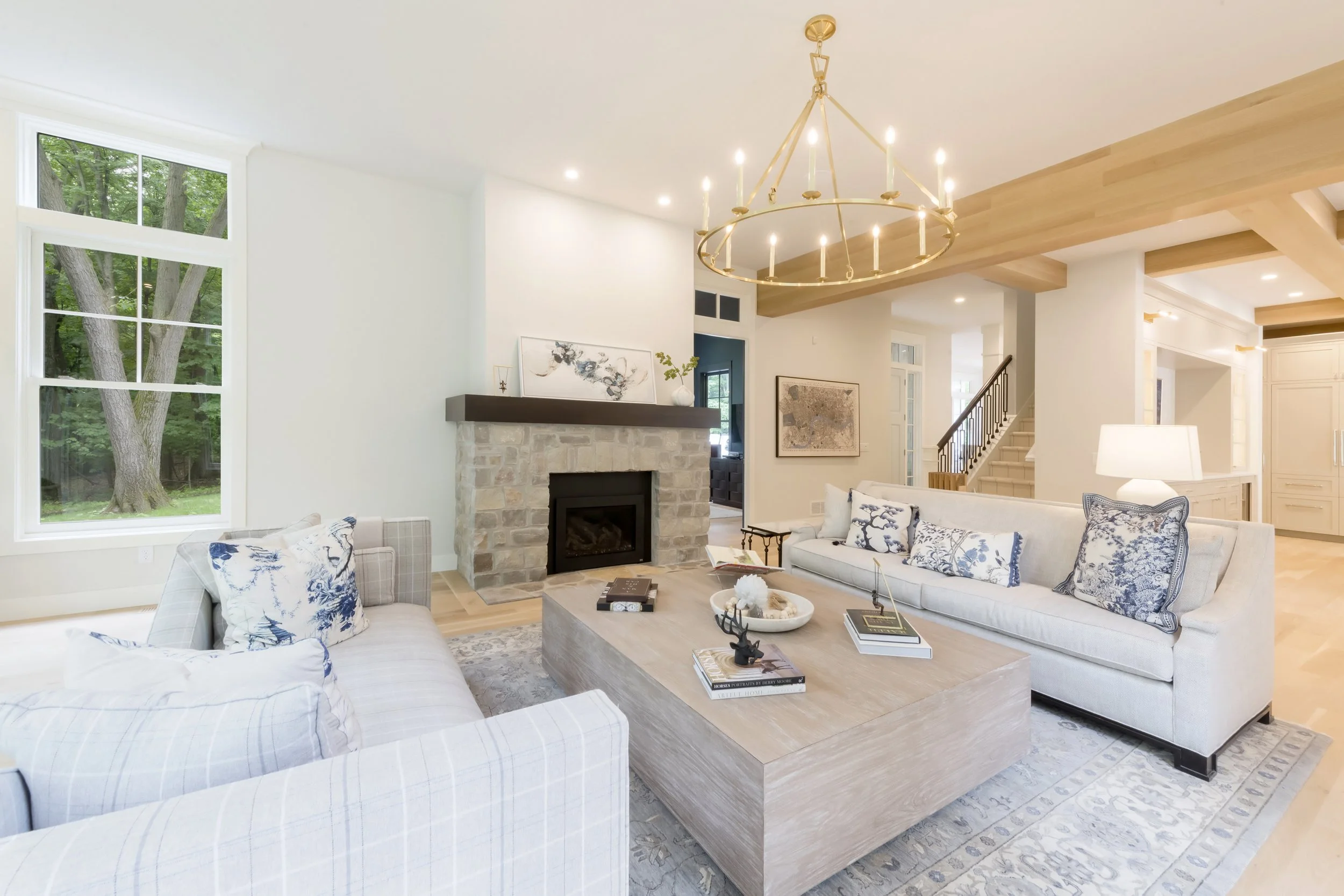 Living room with large window showing outdoor trees, white walls, a stone fireplace, and a gold chandelier. Contains white sectional sofa with blue and white pillows, a wooden coffee table with decorative items, artwork on walls, and staircase in background.