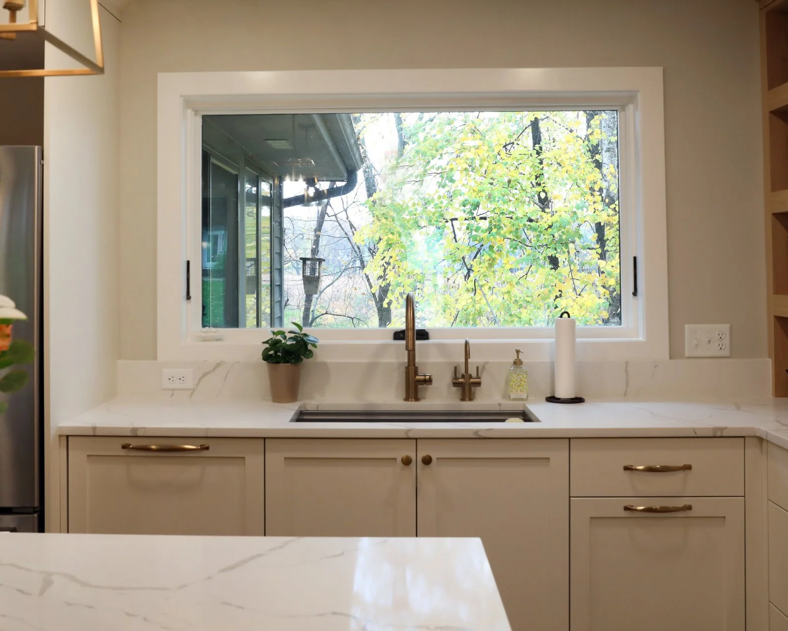 Kitchen with large window showing trees outside, white cabinetry, marble countertop, flowers in a vase, and a refrigerator.