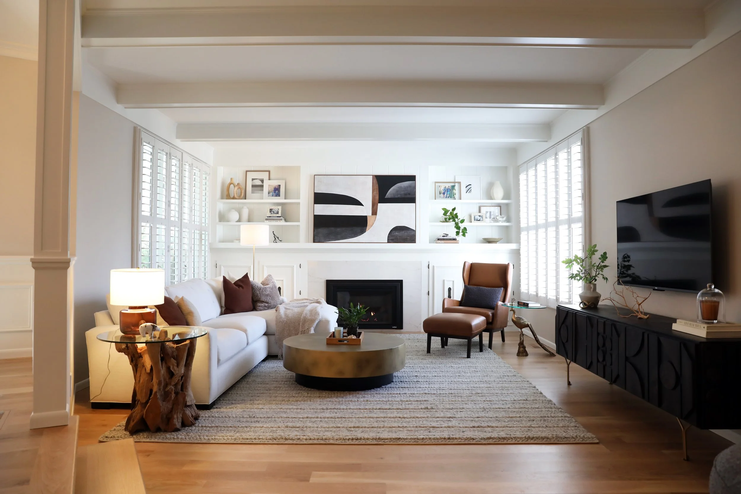 Living room with white sofa, brown armchair, coffee tables, fireplace, wall bookshelf with decorative items and art, large windows with shutters, and flat-screen TV.