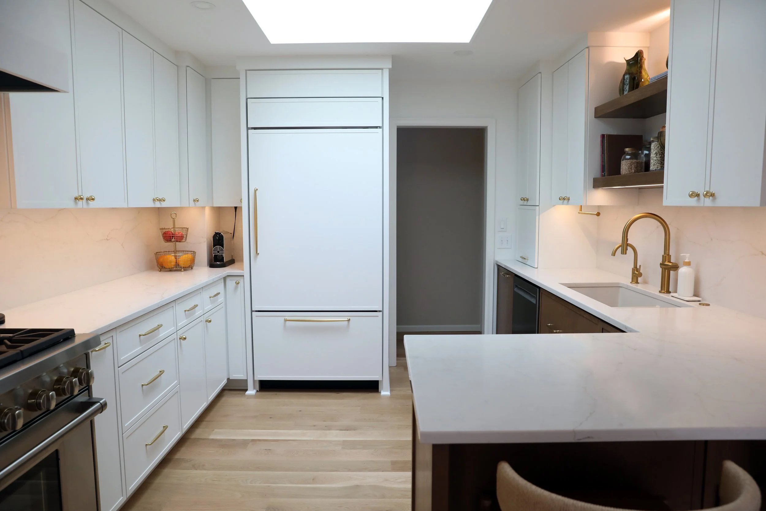Modern kitchen with white cabinets, a white marble countertop, gold hardware, a built-in refrigerator, open shelves with decorative items, a gold kitchen faucet, and a wooden floor.