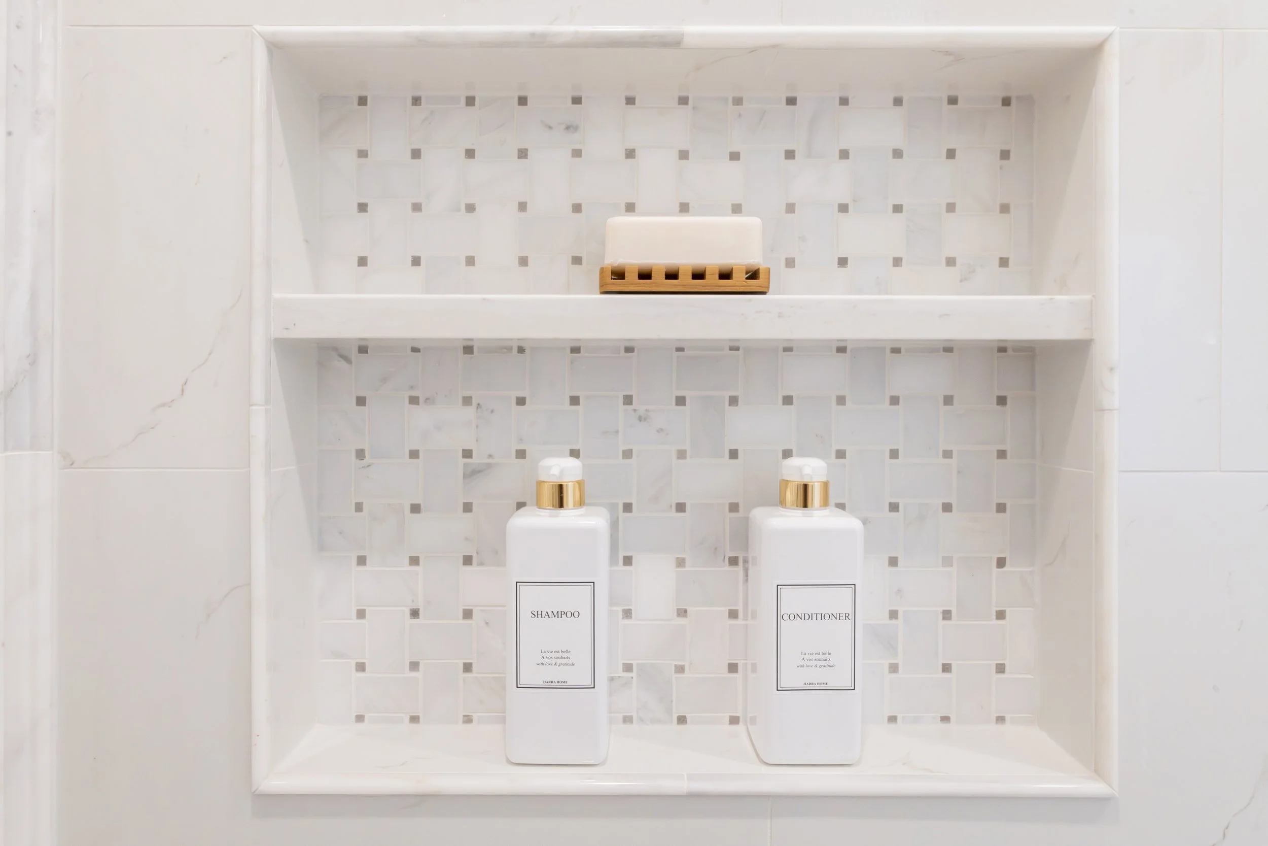 A bathroom shelf with two bottles labeled shampoo and conditioner, a bar of soap on a wooden soap dish, and tiles in the background.