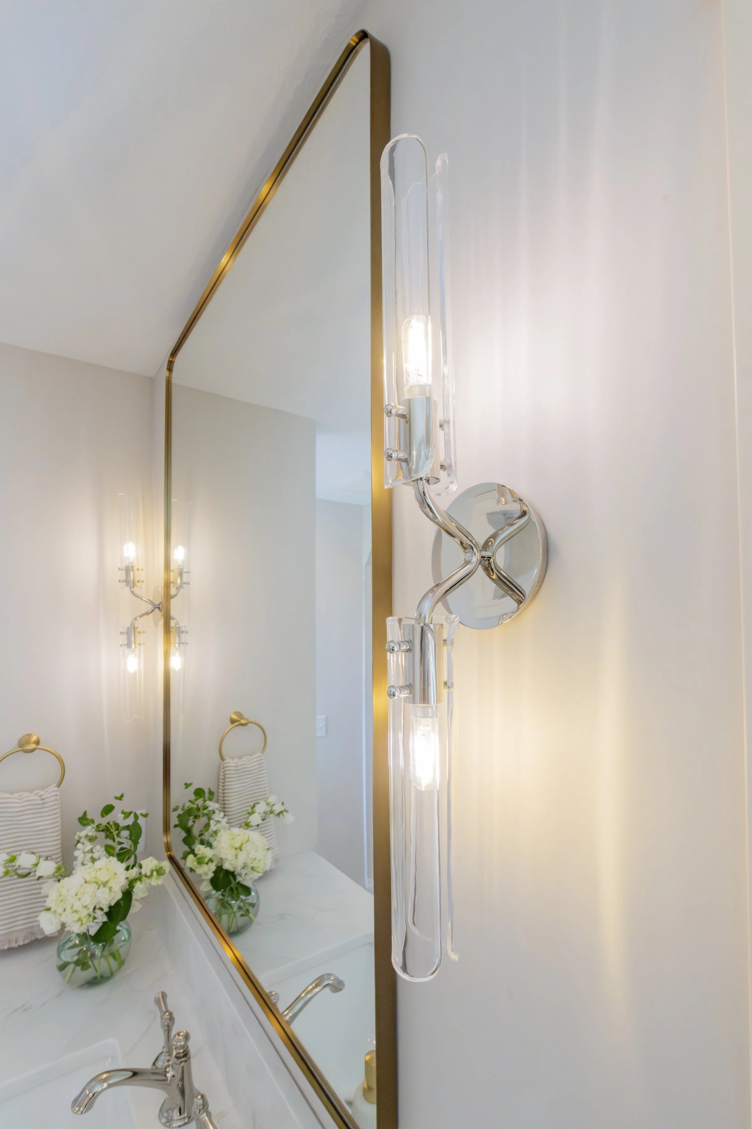 Modern bathroom with gold-accented mirror, wall-mounted light fixture, and white marble countertop. Decor includes glass vases with white flowers, striped towels, and a small green plant.