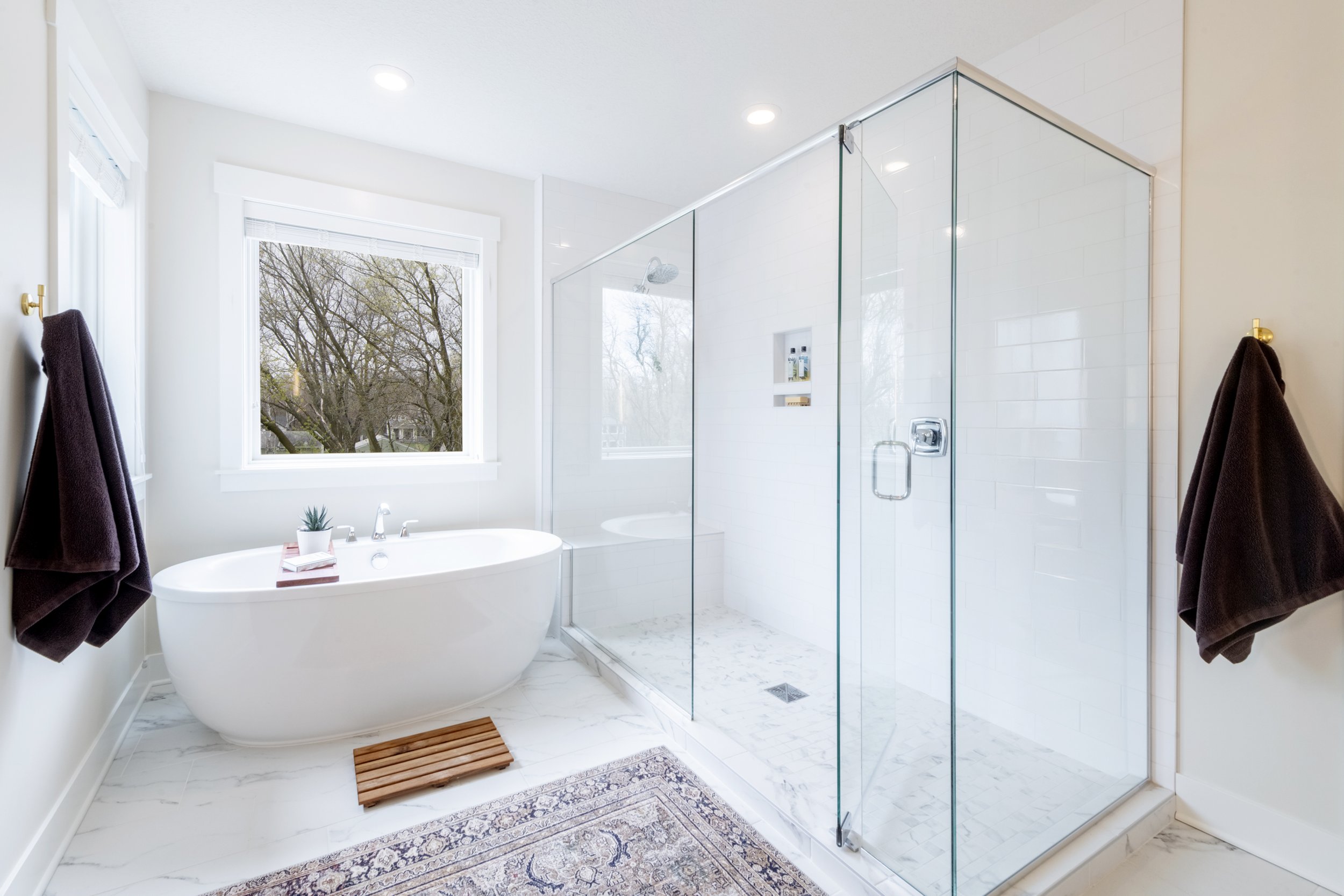 Bright bathroom with a large window showing trees outside, a bathtub, and a glass-enclosed shower with white tiles.