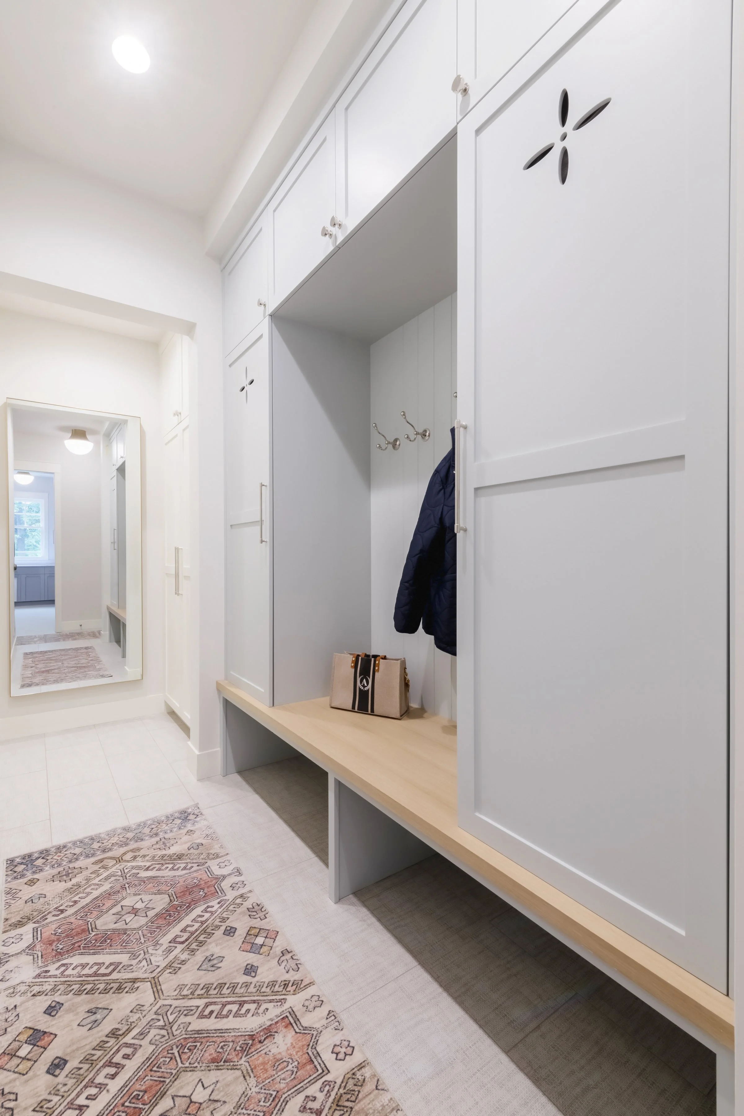 A hallway with white built-in cabinets and a wooden bench; mirrors on the wall, coat hooks with a dark jacket hanging, beige and patterned area rugs on a white tiled floor, with a door and window visible in the background.