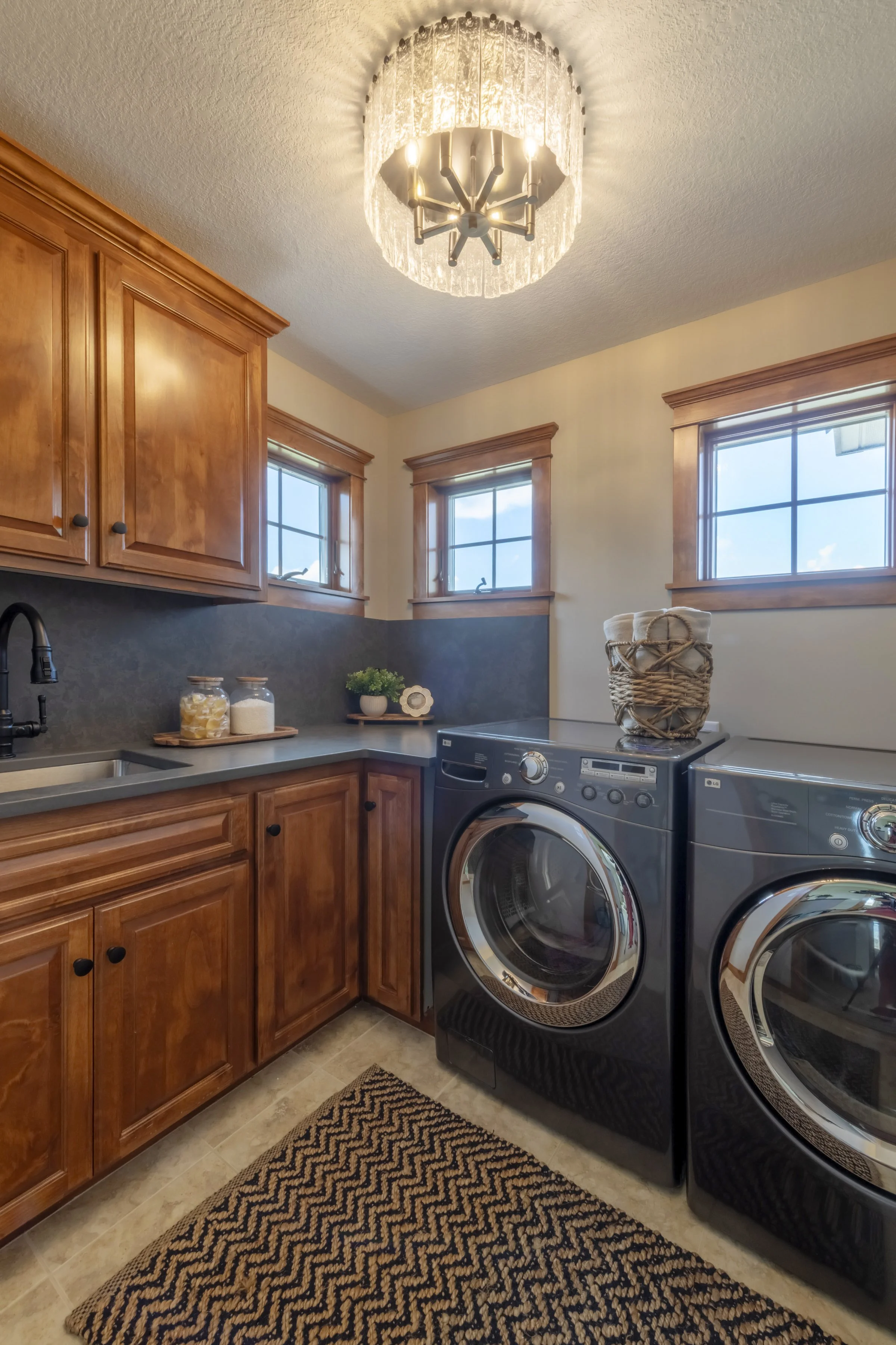 A laundry room with wooden cabinets, two windows, and a front-loading washing machine and dryer. There is a woven basket on top of the washer, and a patterned rug on the floor.