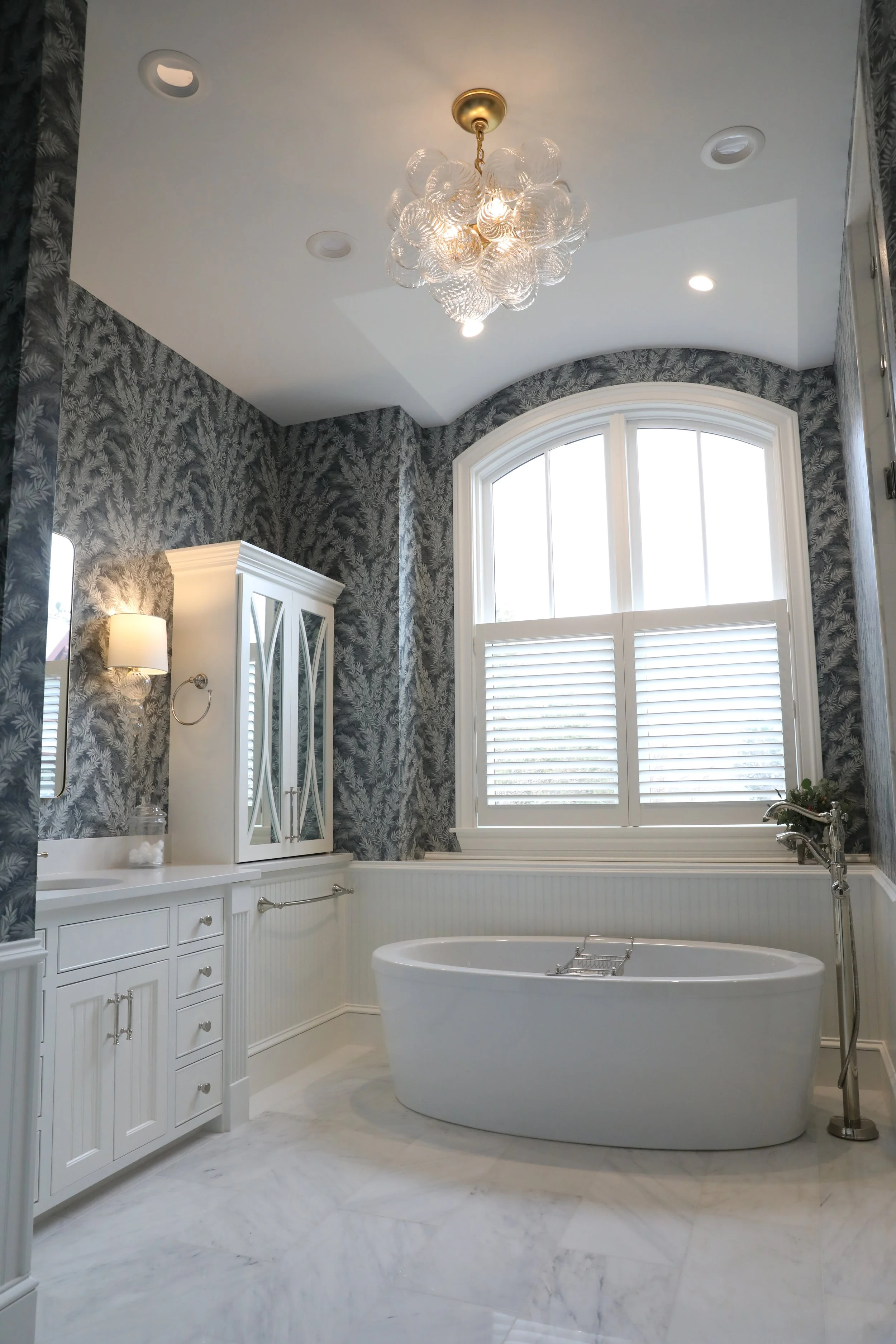 A luxurious bathroom featuring a freestanding bathtub, a white cabinet with a mirror, and a large arched window with white plantation shutters. The ceiling has a modern glass bubble chandelier, and the walls are decorated with dark, leafy patterned wallpaper. Marble flooring completes the elegant look.