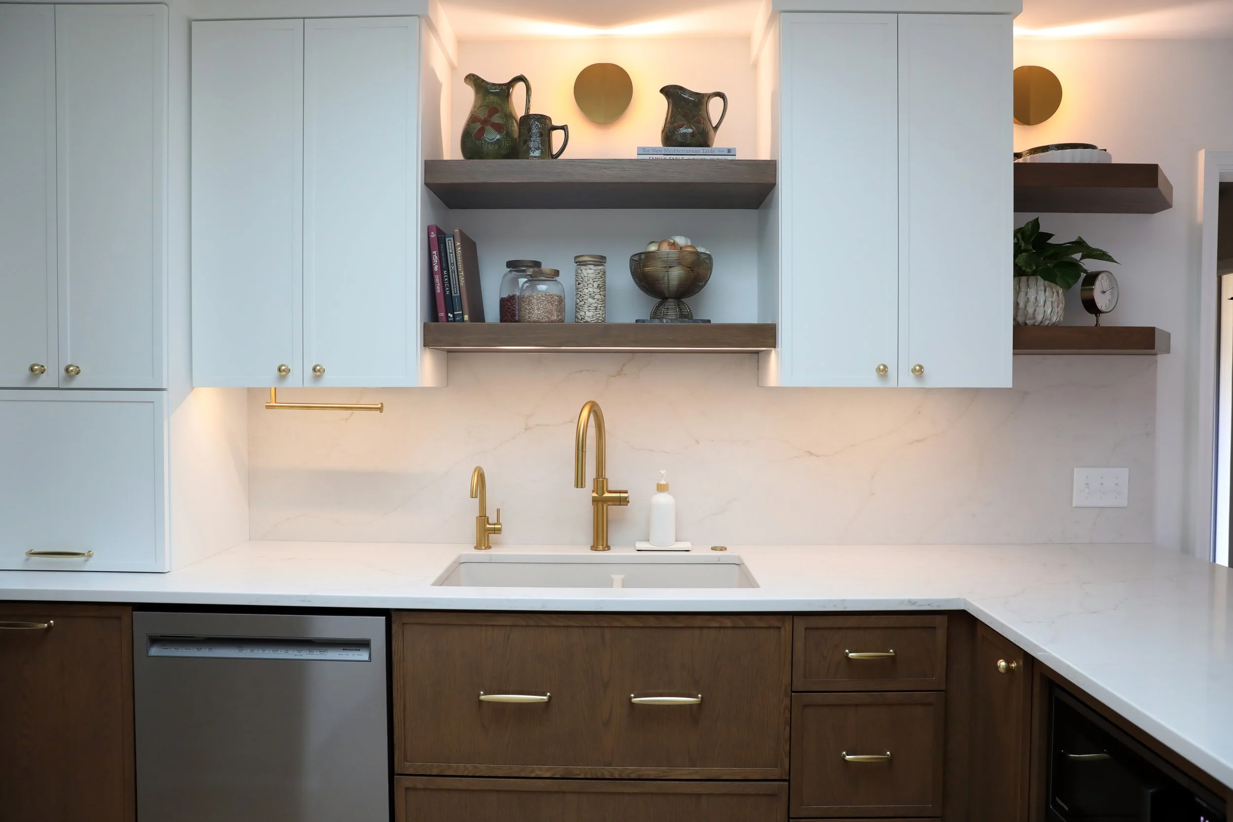 Modern kitchen with white upper cabinets and dark lower cabinets, featuring brass handles and a gold faucet, marble backsplash, open shelves with decorative items, jars, books, and a plant, with a section of the sink and dishwasher visible.