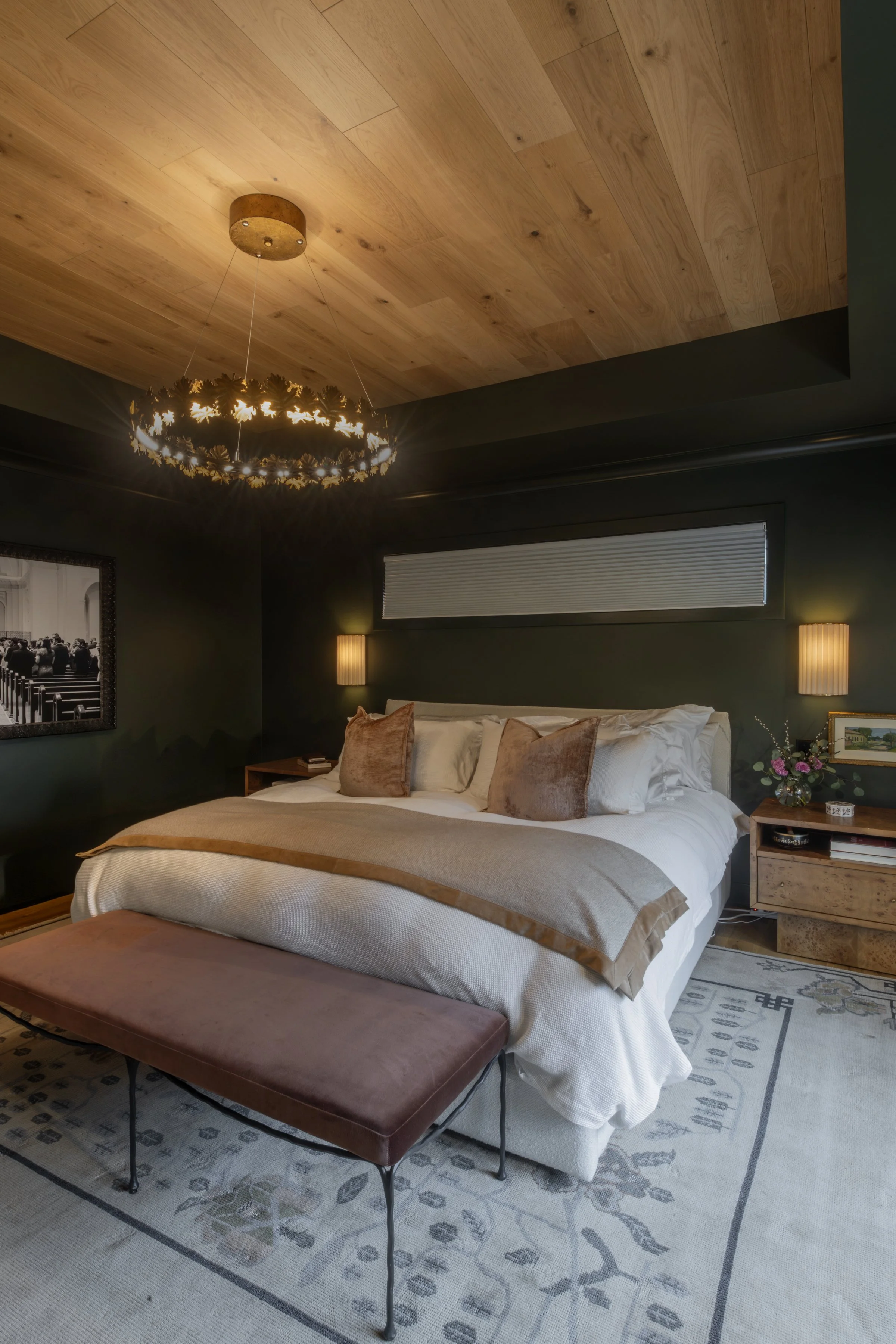 A bedroom with a bed, throw pillows, a nightstand with a flower arrangement, black wall, window with blinds, and a decorative chandelier hanging from a wooden ceiling.