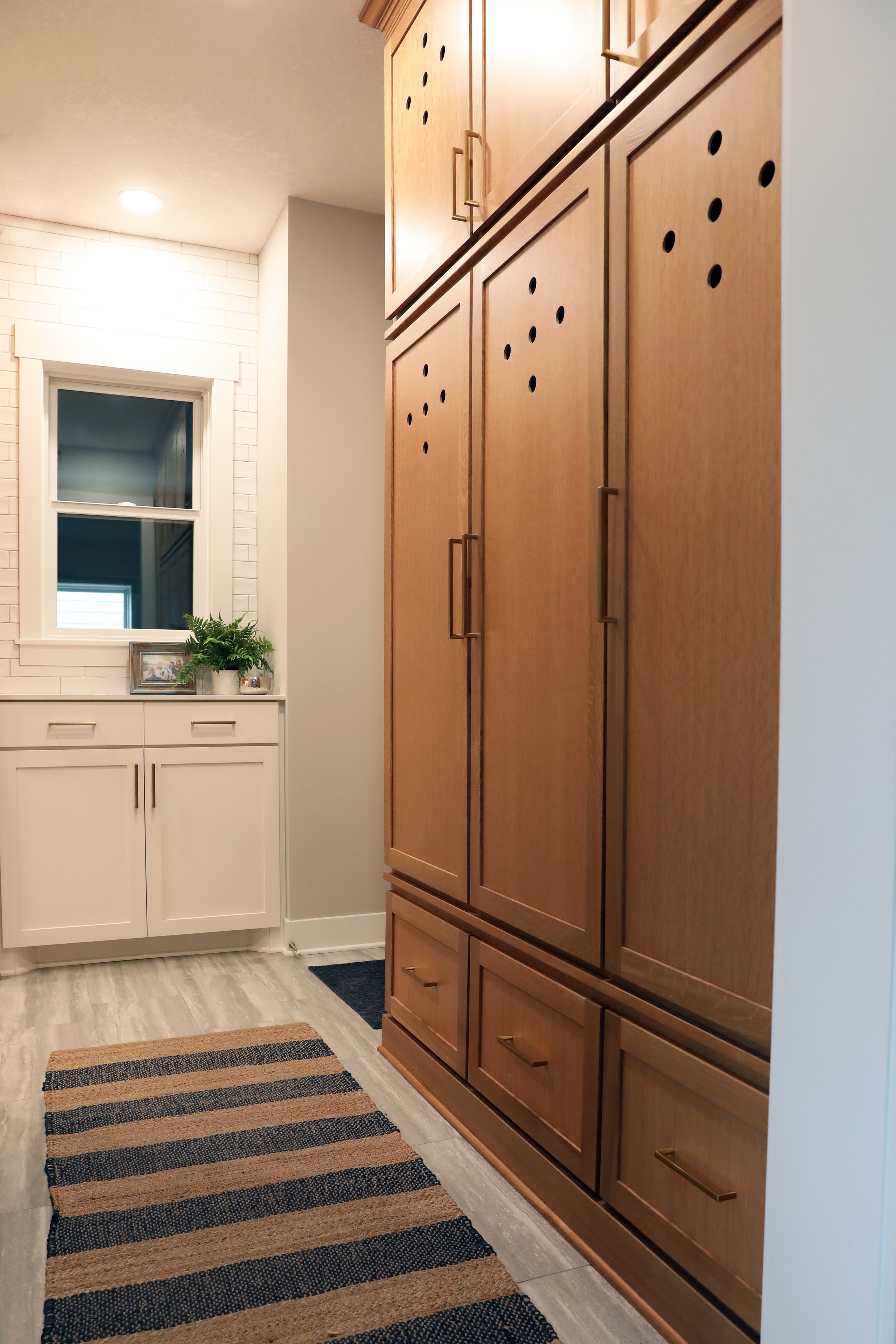 Interior of a home hallway featuring a large wooden cabinet on the right with multiple doors, a white cabinet with drawers and a small framed photo and potted plant on top, a window with white trim and brick accent wall, a striped rug on the light-co
