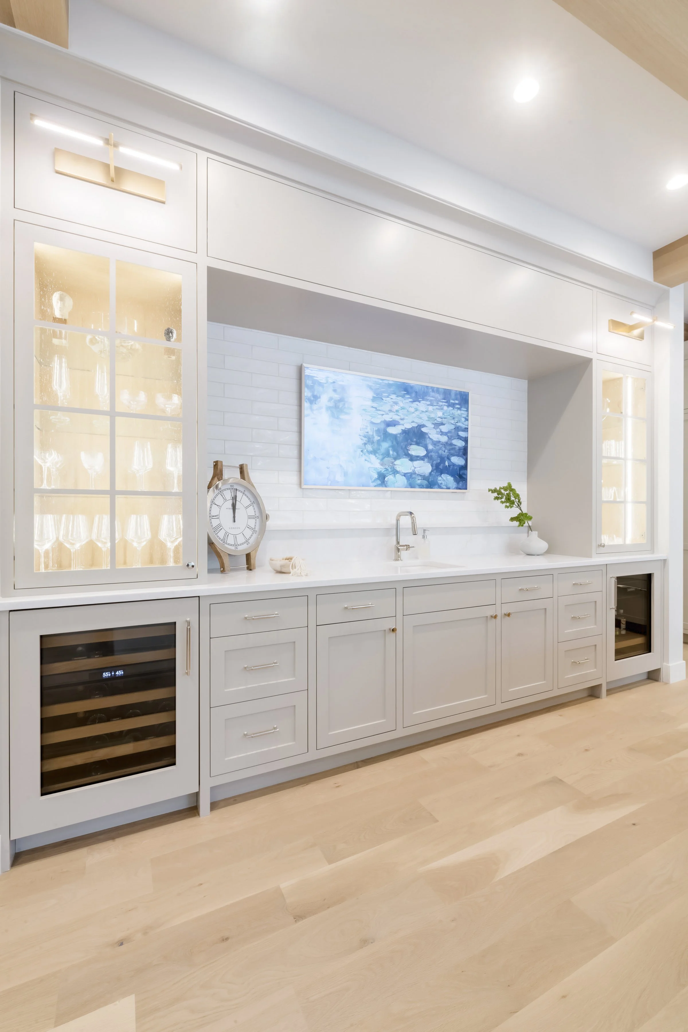 Modern white kitchen with a built-in wine fridge, a counter with a clock, a sink, a small potted plant, and a colorful pond picture on the wall.