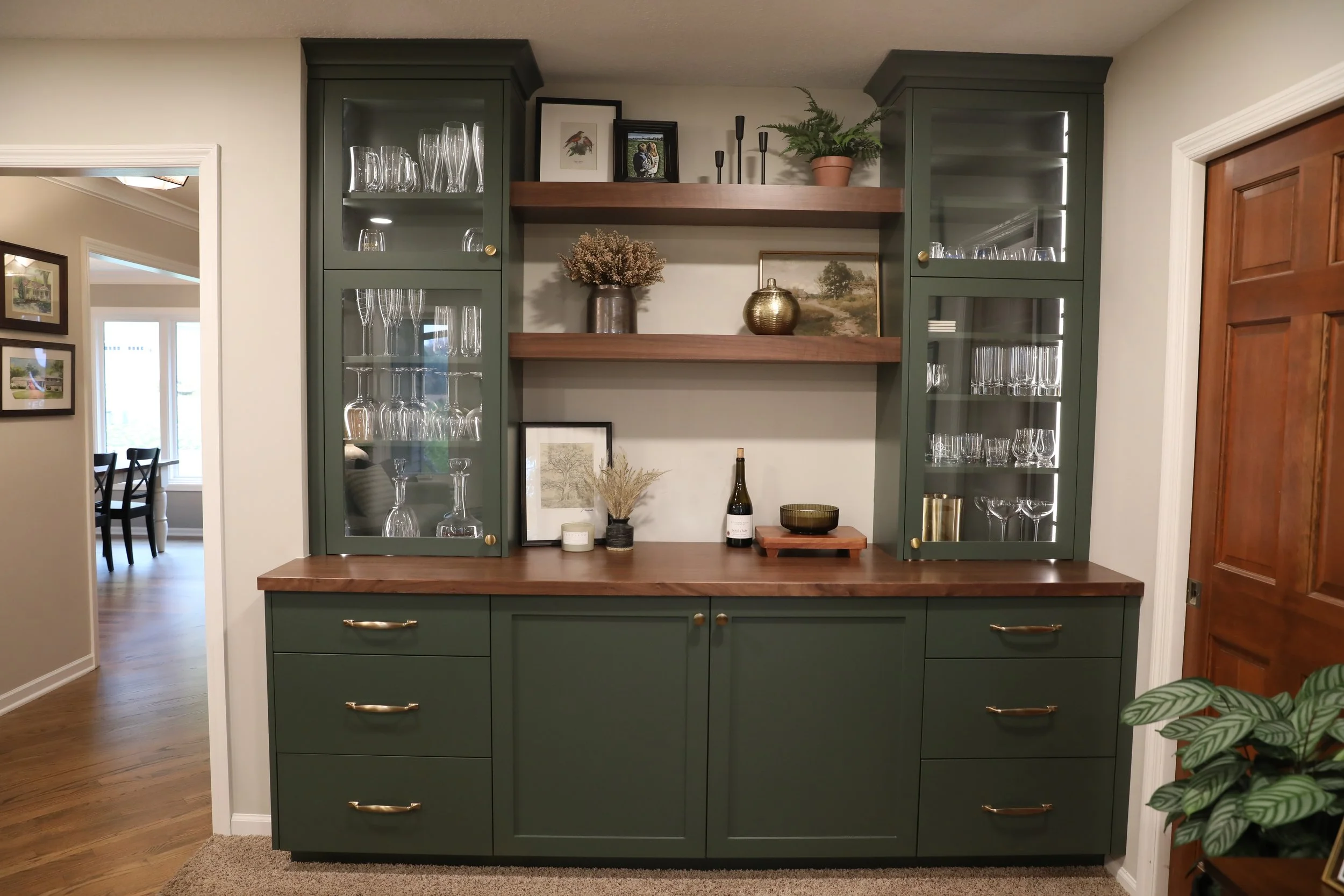 A green kitchen cabinet with glass doors displaying glassware, with decorative items including framed pictures, a potted fern, and a gold vase on wooden shelves above.