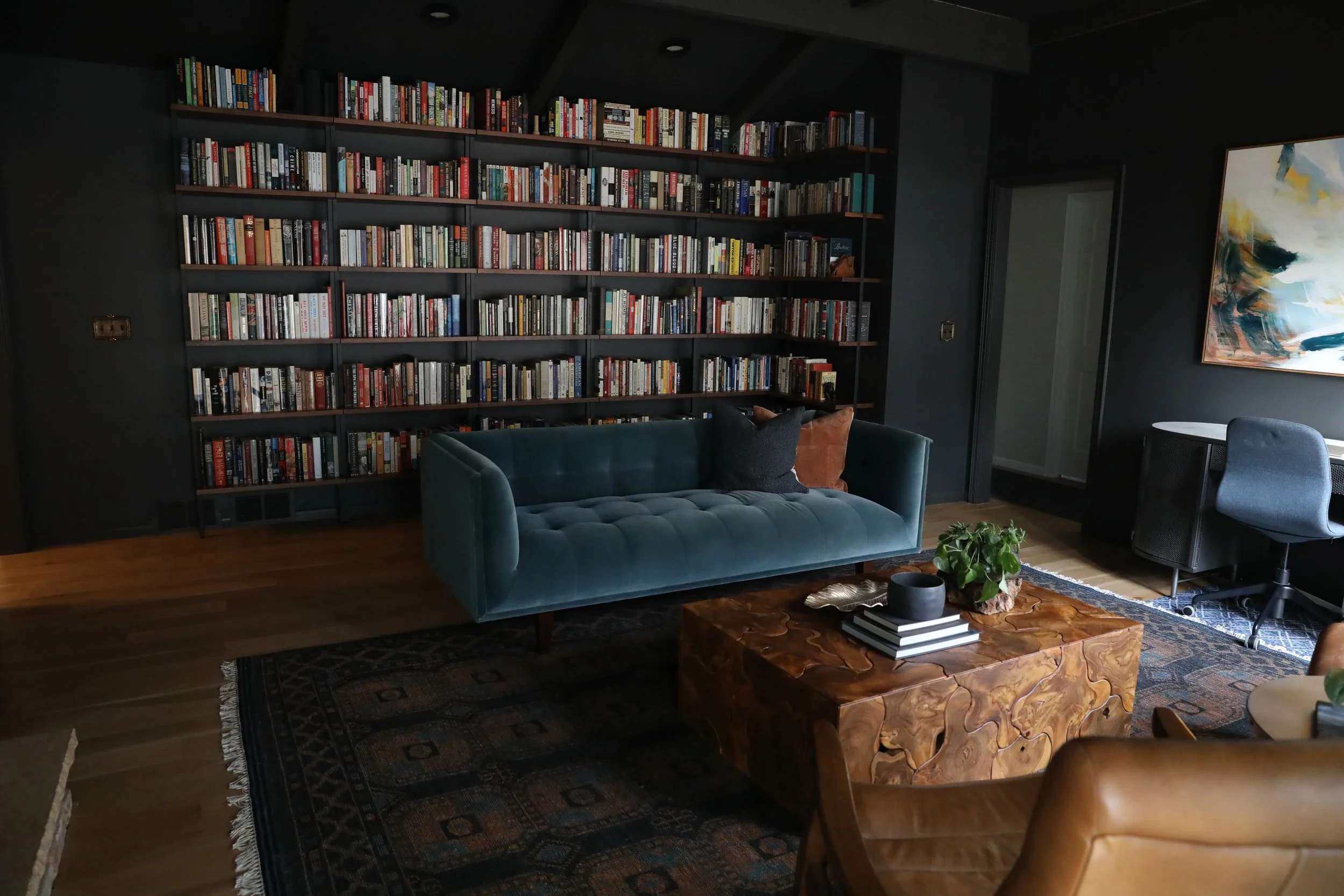 A dark-colored room featuring a large bookshelf filled with books, a green velvet sofa with pillows, a wooden coffee table with a plant and books, a patterned rug, a blue chair, and a colorful abstract painting on the wall.