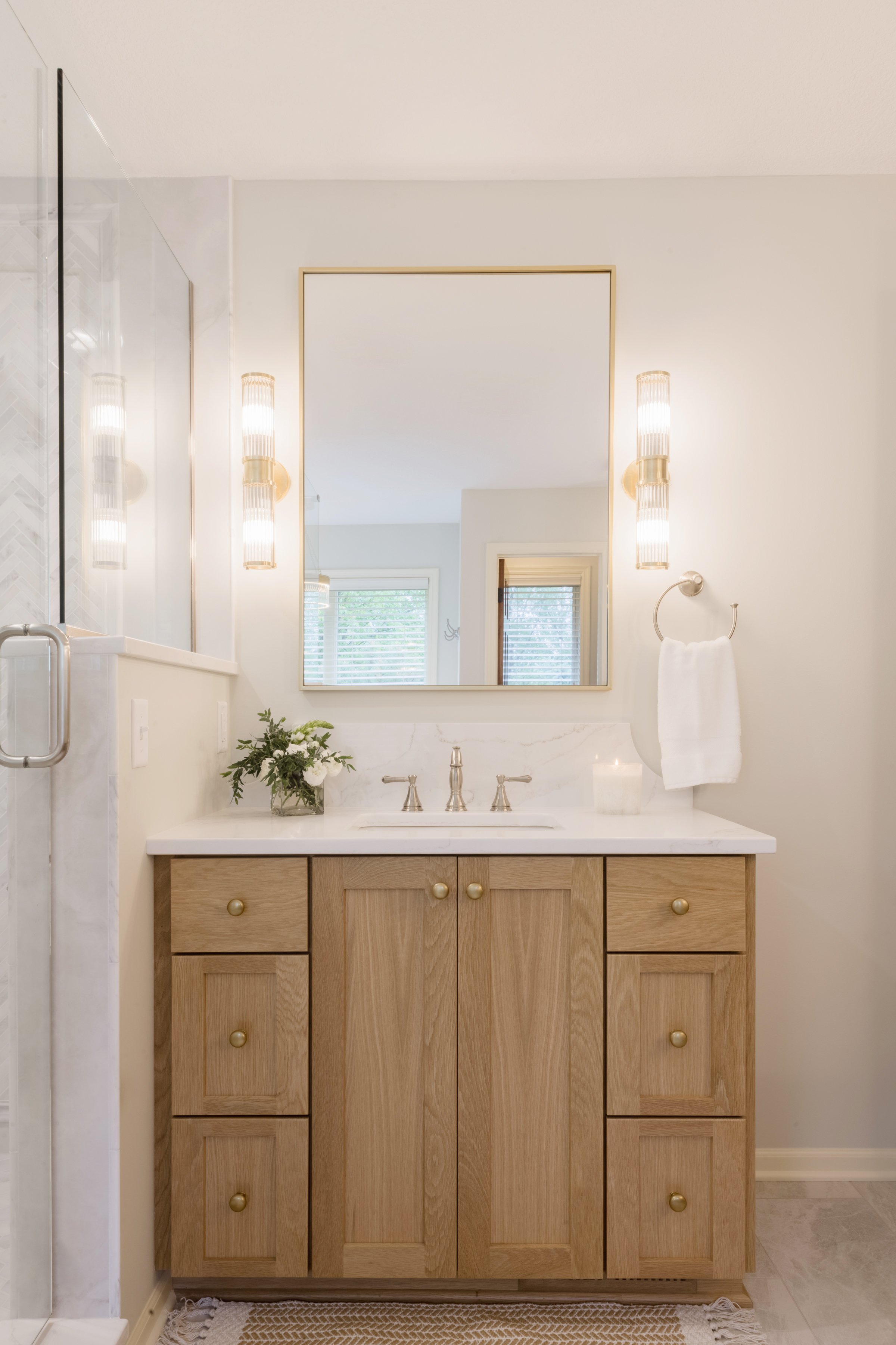 Bathroom vanity with a wooden cabinet, white marble countertop, and a large mirror. Two modern vertical light fixtures are mounted on either side of the mirror, and a small plant and a candle are placed on the countertop. A towel hangs on a hook next