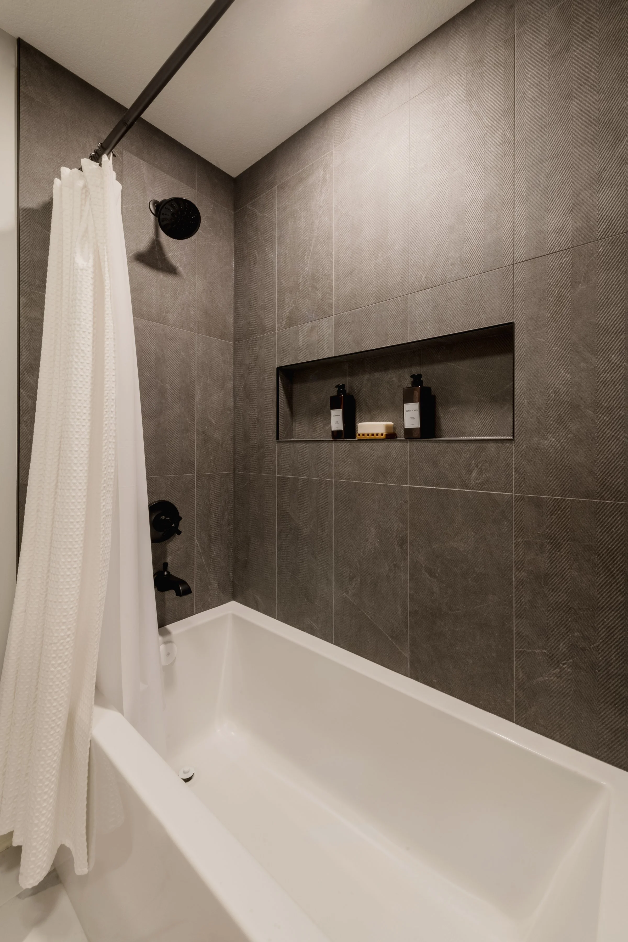 A modern bathroom with brown textured tile walls, a white bathtub, a black showerhead, and a built-in wall niche with bottles and a soap dish. White shower curtain partially visible.