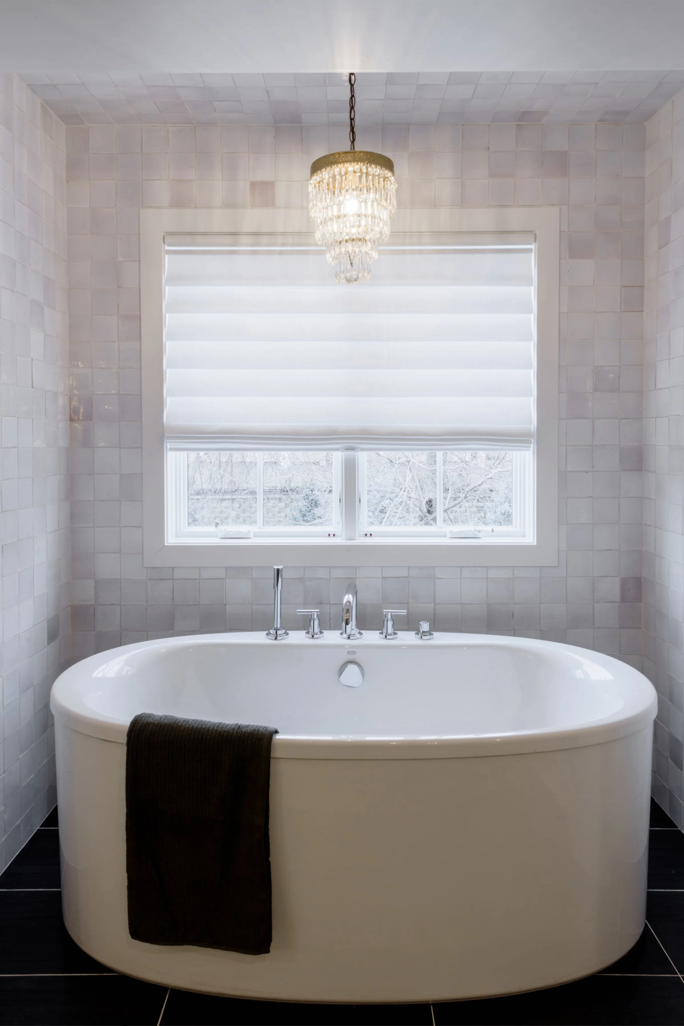 A white free-standing bathtub with a black towel draped over the side, set against a wall with small white tiles. Above the tub, there is a window with white blinds, and hanging from the ceiling is a decorative chandelier with crystal accents.