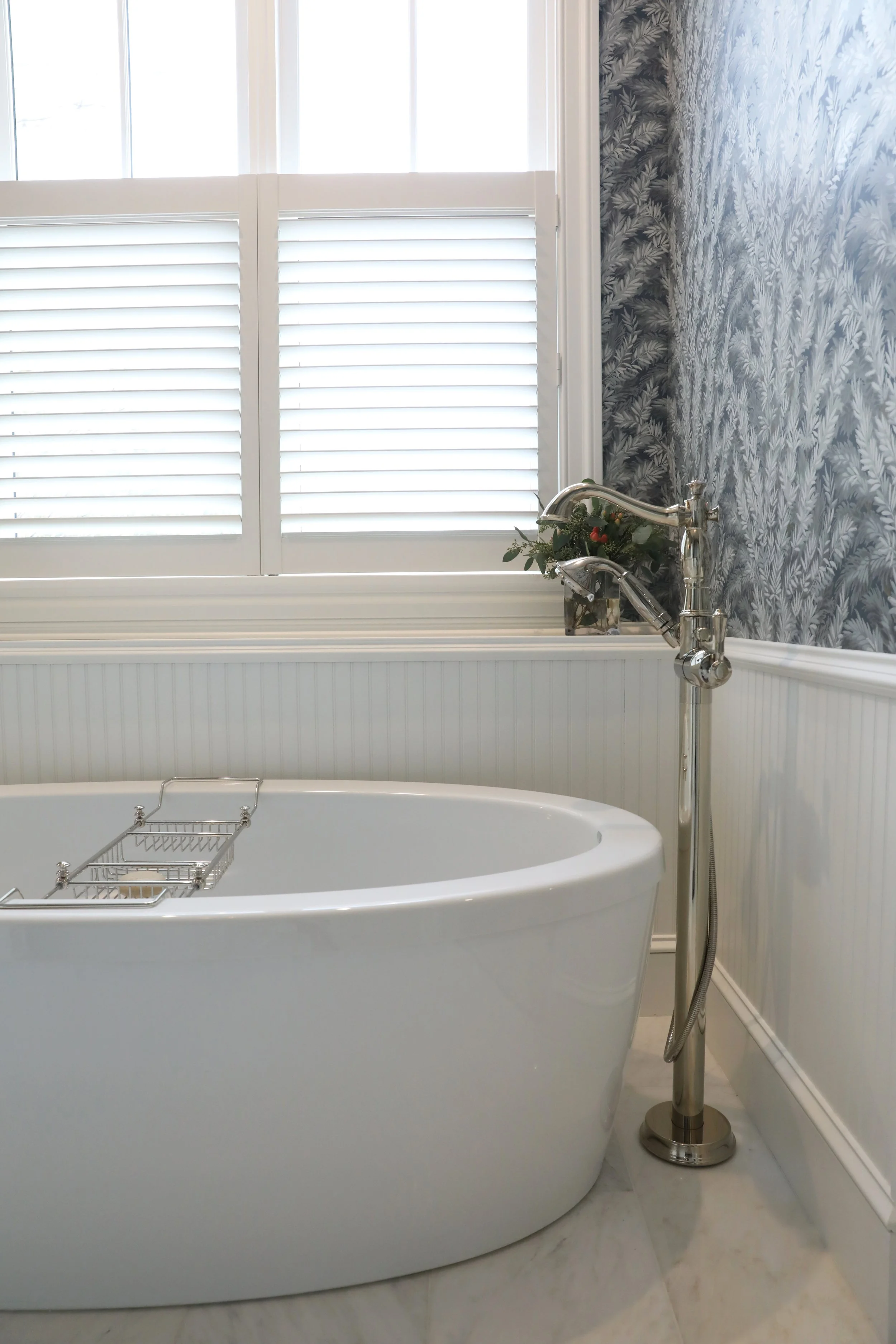 A white bathtub with a metal rack inside, positioned next to a silver bathtub faucet and a standing silver faucet with a sprayer, in front of a window with white plantation shutters and a wall with a gray leafy pattern wallpaper.