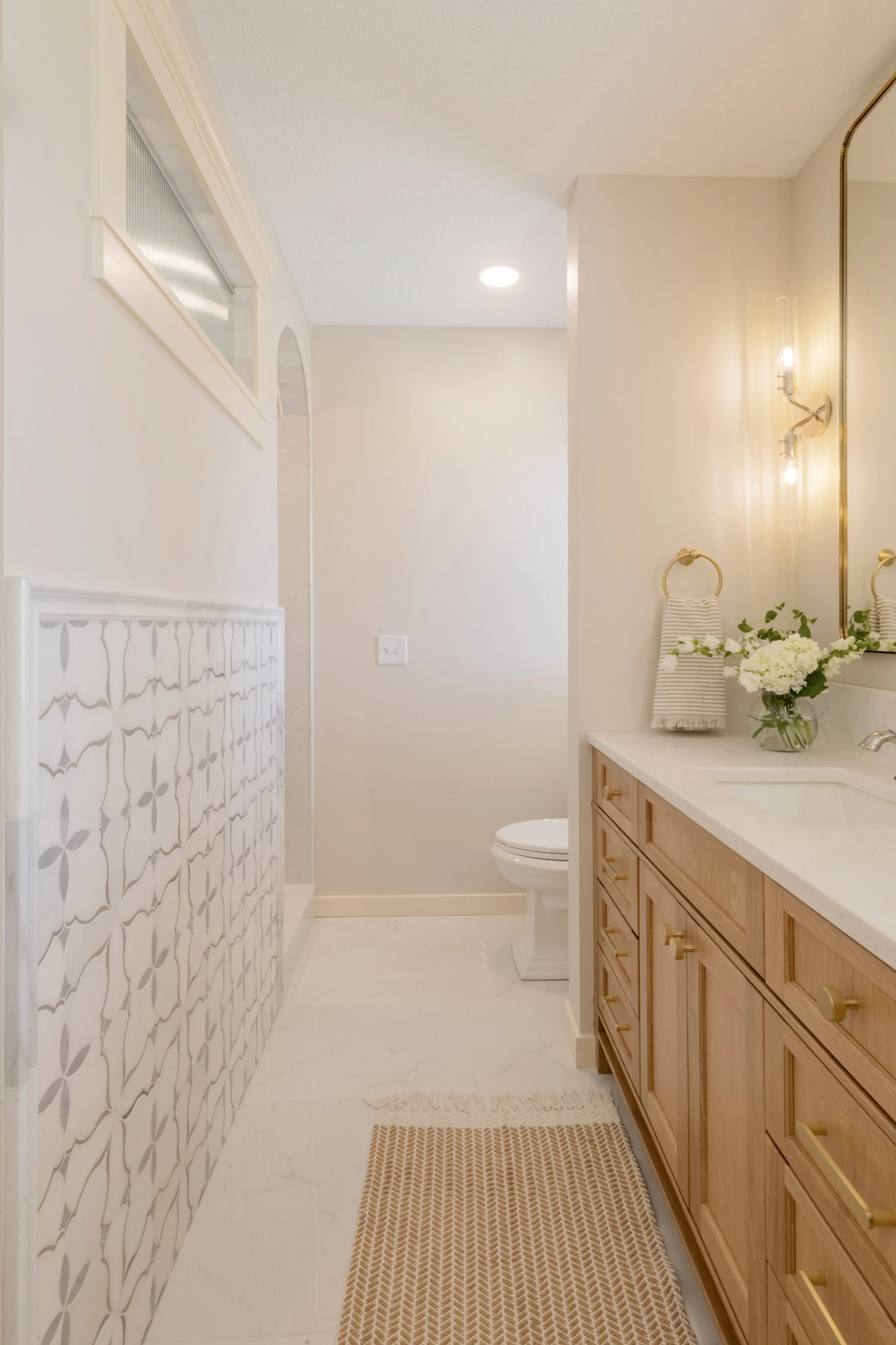 A bathroom with a light wood vanity, white countertop, and mirror. There is a white toilet, wall-mounted light fixtures, and a towel rack with a striped towel. A vase with flowers is on the countertop. The walls are painted in a neutral color, and th
